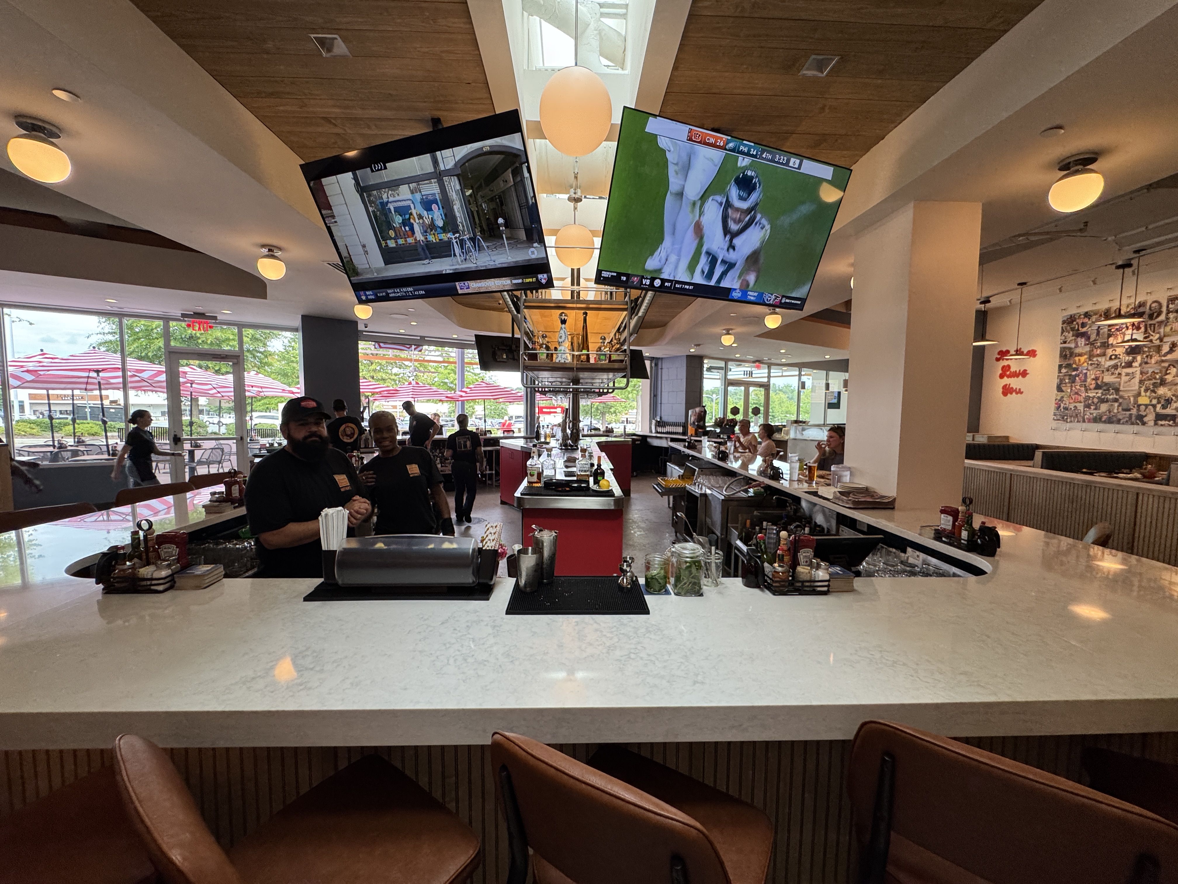 Interior of a modern bar with a U-shaped white marble counter, brown stools, two TVs overhead showing a football game and a street view, bartenders behind the counter, and red umbrellas outside.