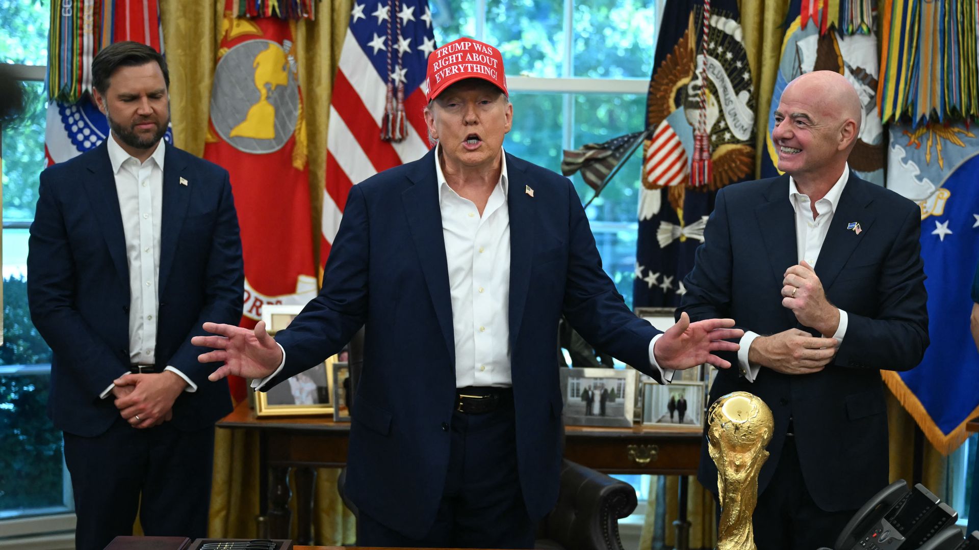 Photo of three men behind a desk with a gold trophy on it. The man in middle is wearing a red hat.