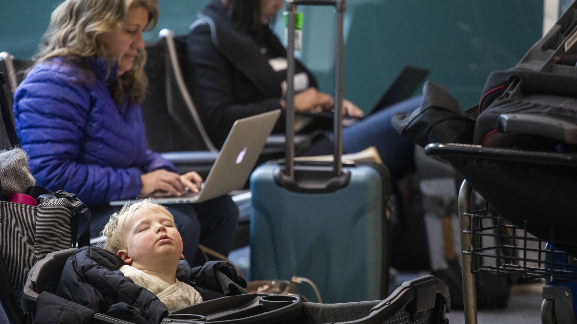 A child in the foreground naps while inside a stroller, while a woman on a laptop in the background studies her screen. Besides them are luggage and another person sitting in the background. 