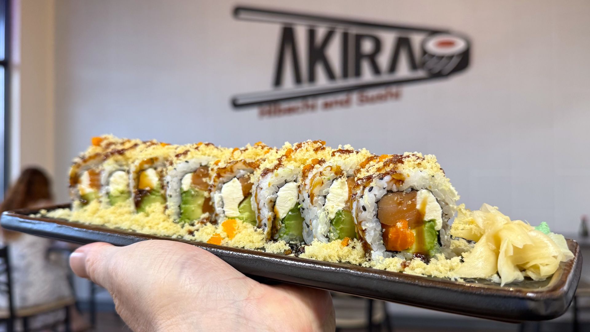 A hand holds a sushi roll topped with tempura flakes and sauce at Akira Sushi, with ginger and wasabi on the side.