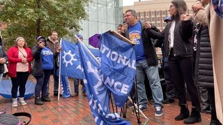 TPS holder Jose Palma, center, holds hands with Boston City Councilor Gabriela Coletta  as he stands behind a podium.