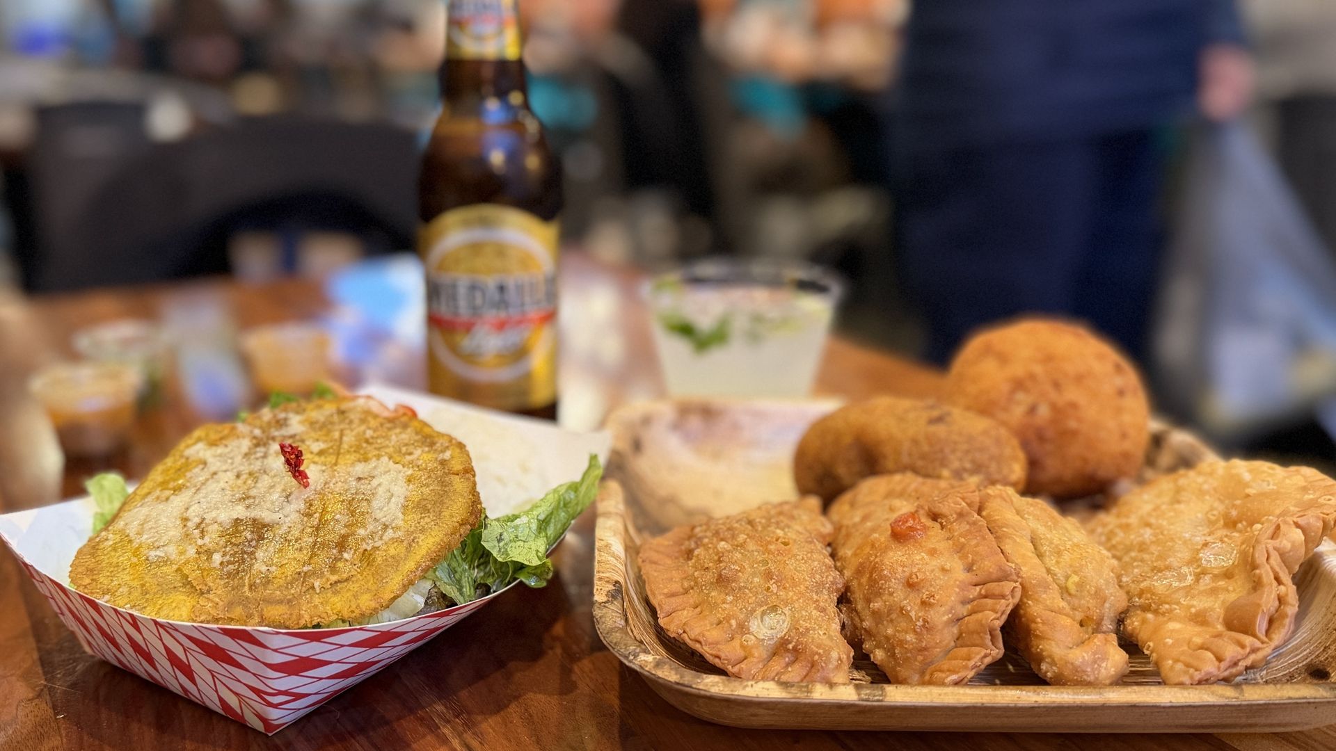 Close-up of party food on a table: a sandwich with greens in a red-and-white striped paper basket, a beer bottle behind, and a tray of fried pastries to the right; blurred people in the background.