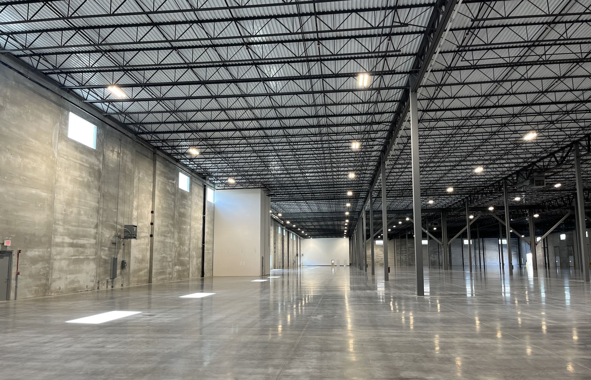 Interior of a large empty industrial warehouse: gray concrete walls, windows along the left wall, a black metal truss ceiling with bright overhead lights, tall columns, and a polished concrete floor.