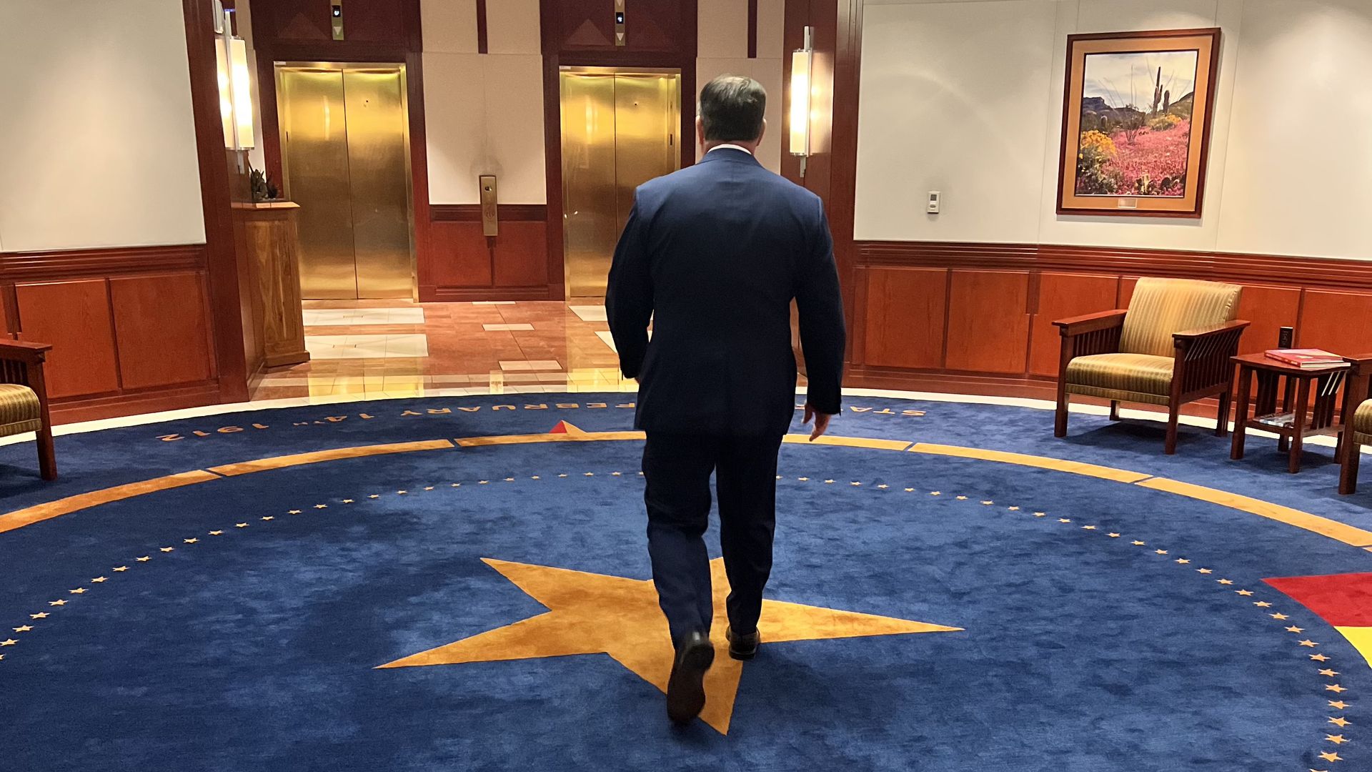 Doug Ducey's back faces the camera as he walks across an Arizona flag carpet toward elevator doors