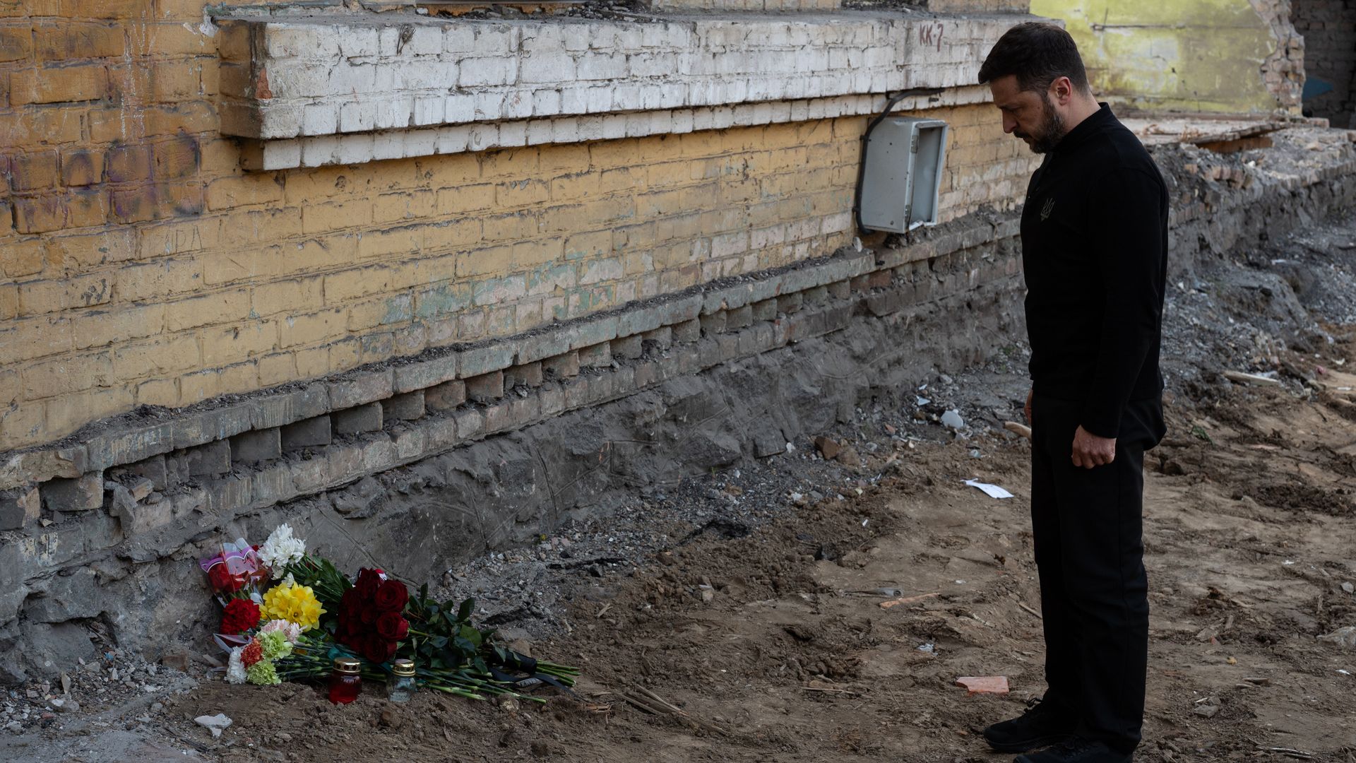 A short, dark-haired, bearded Ukrainian President Volodymyr Zelensky, wearing a black jacket and pants, bows his head toward a floral tribute at the site of a Russian rocket attack that occurred on April 24.