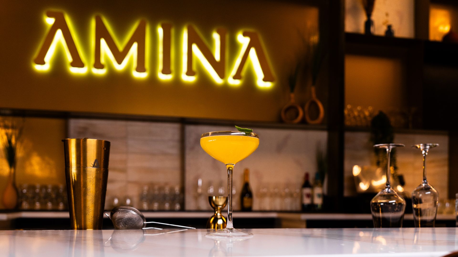 Cocktail glass with bright orange drink garnished with a leaf on a marble bar counter, with bar tools and illuminated "AMINA" sign in warm yellow light in the background.