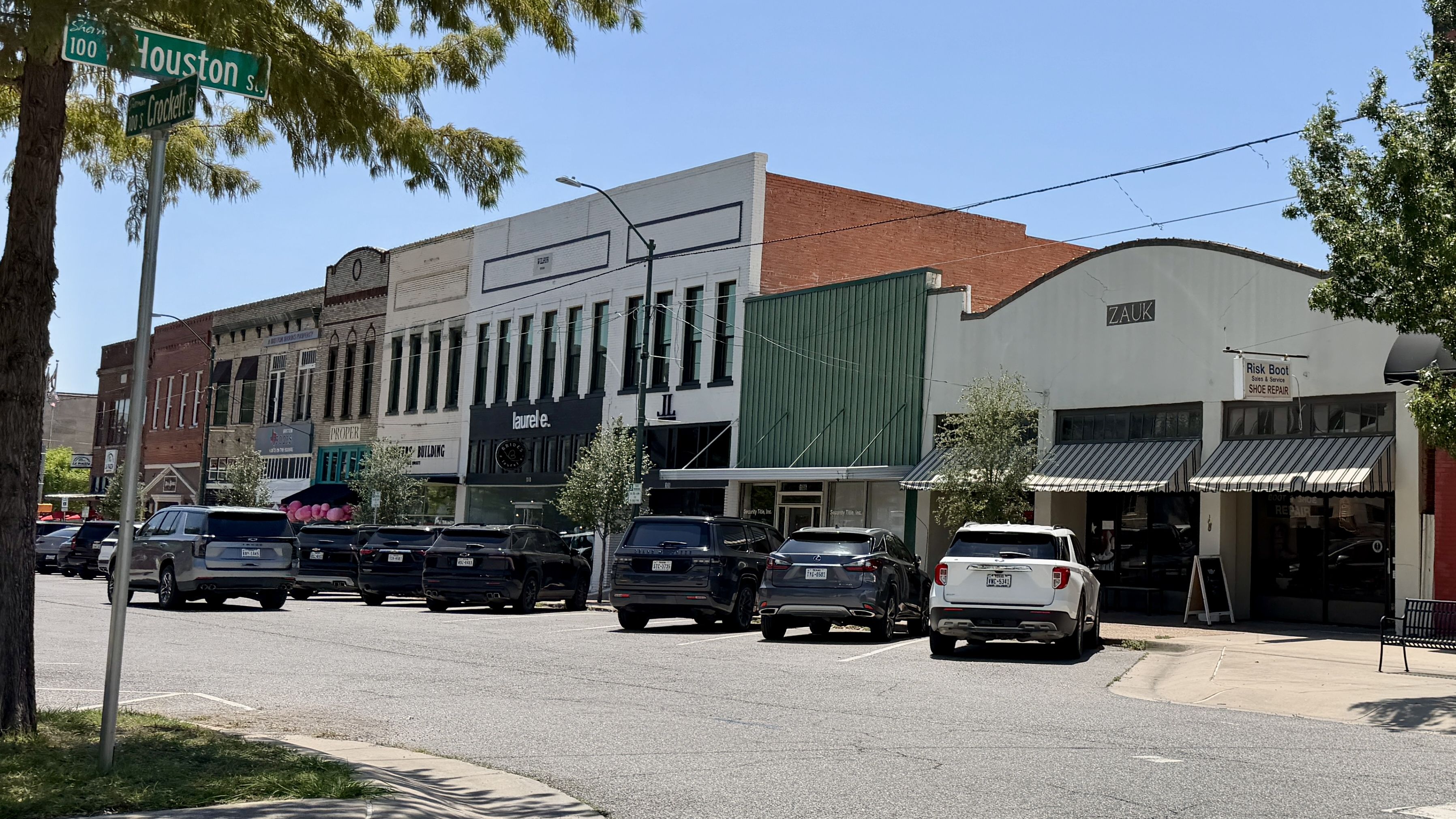 Sunny downtown street with a row of storefronts, brick and white façades, a green metal wall, and parked cars along the curb. Green street signs mark the intersection at Houston Street.