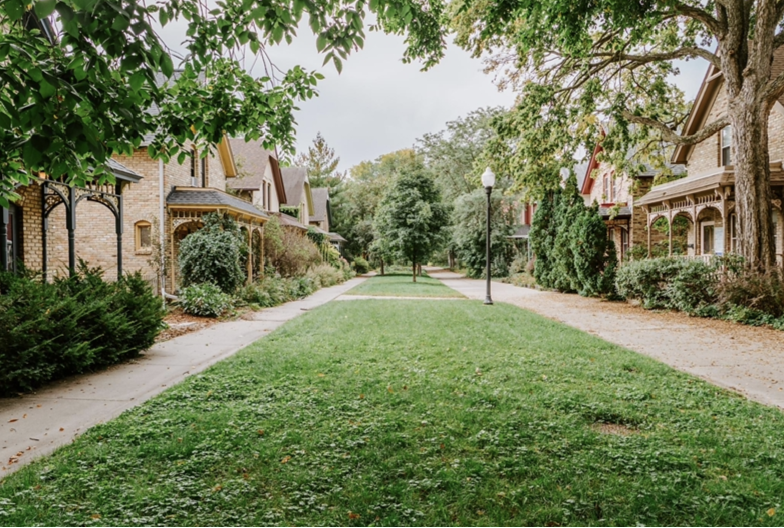 Green grassy path flanked by sidewalks and rows of brick houses with wooden porches shaded by leafy trees under a cloudy sky.
