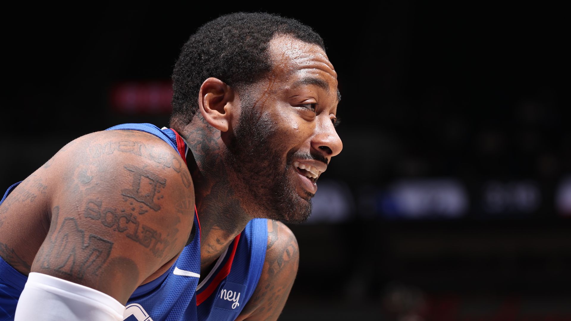 Close-up of a smiling basketball player with tattoos on his shoulder and neck, wearing a blue jersey with red and white trim, against a dark blurred background.