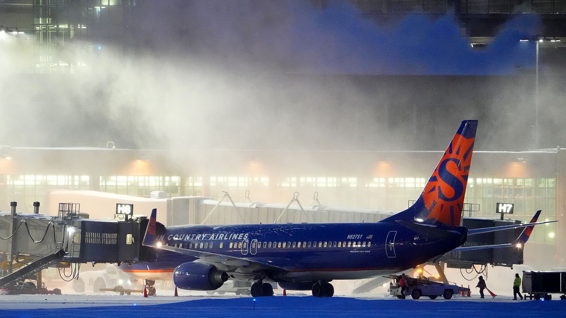 A blue Sun Country Airlines plane on the tarmac near an airport jetbridge with snow falling around it.