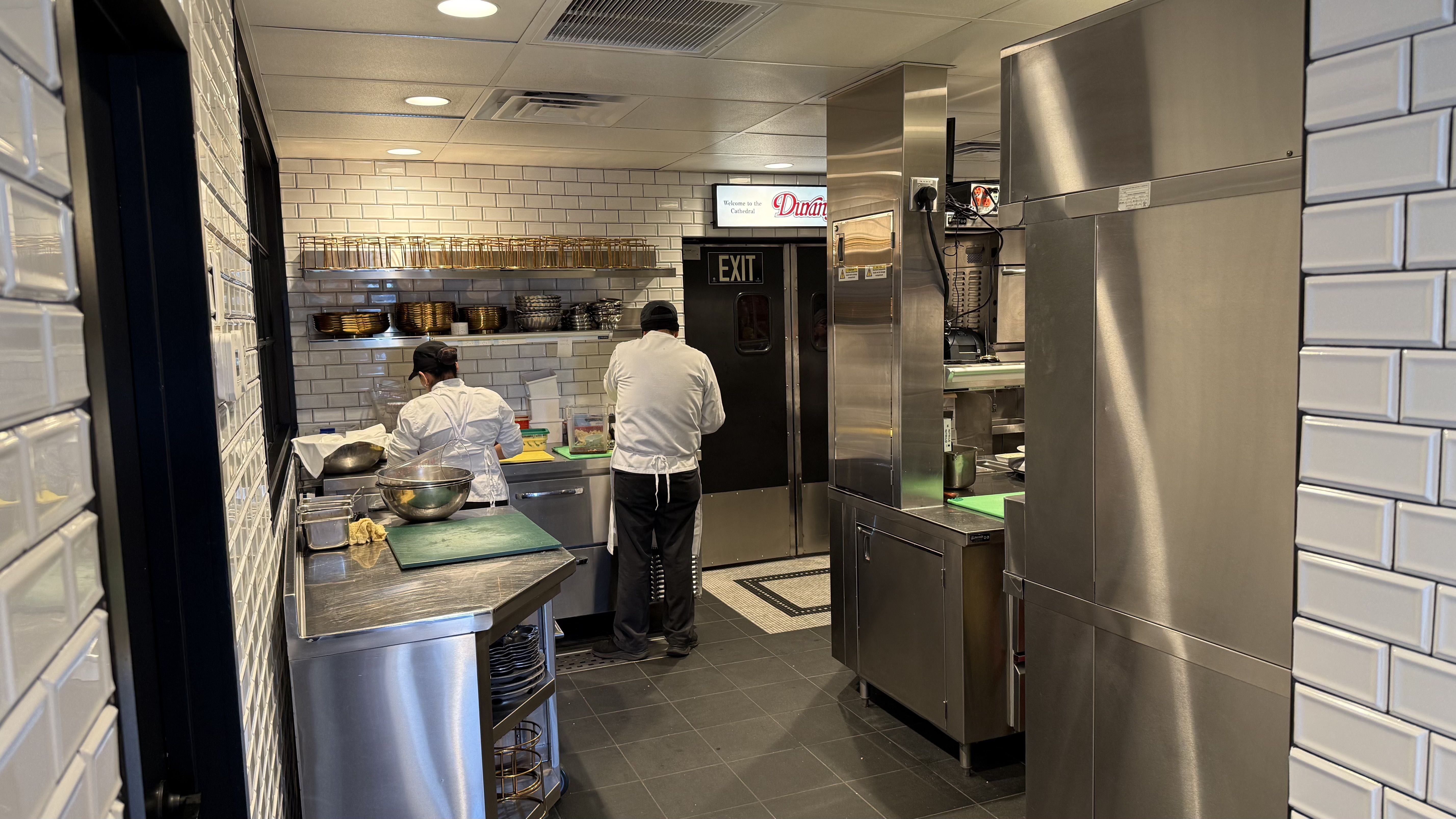 Two chefs in white uniforms working in a stainless steel kitchen with white tile walls, shelves of bowls and glasses, and a black EXIT door with a small lit sign above it.
