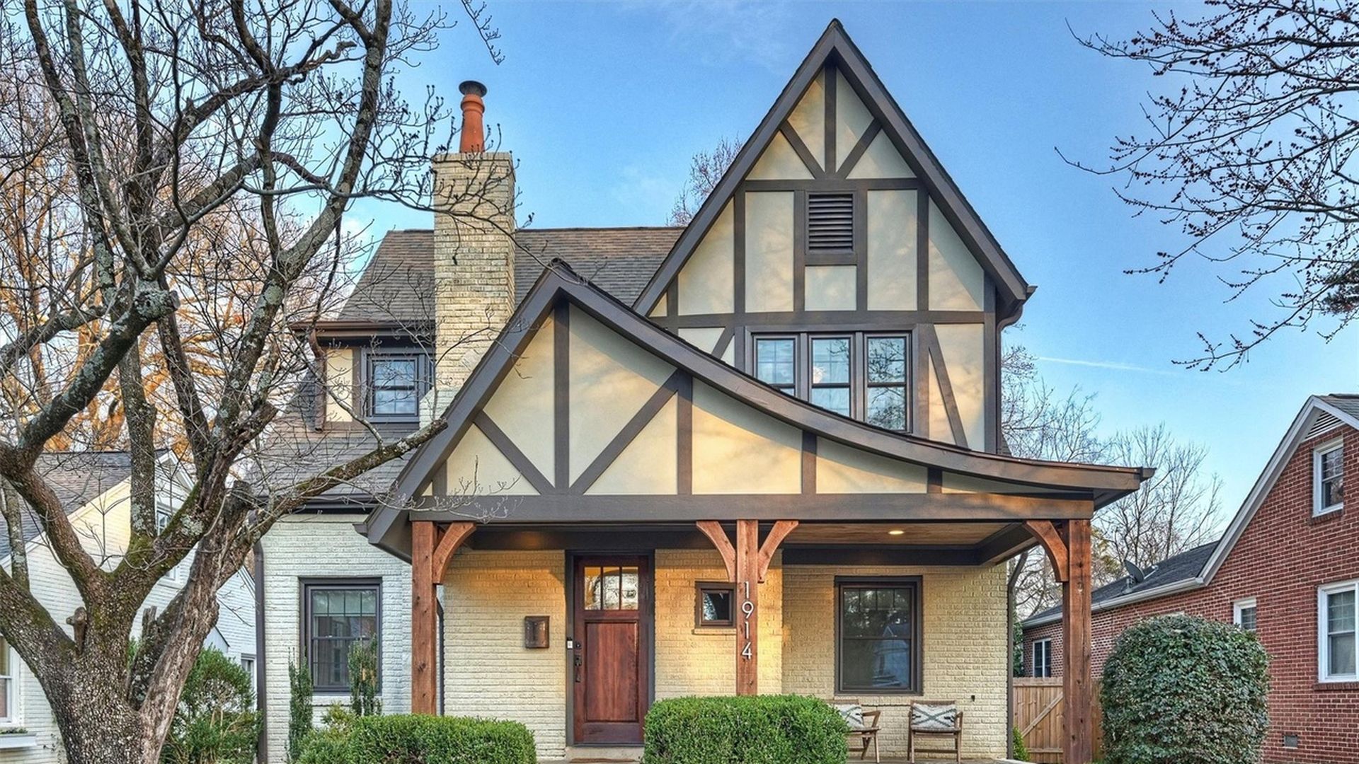 Two-story Tudor-style house with beige and brown exterior, wooden porch columns, a chimney, and a front lawn with trimmed bushes and a leafless tree under a clear blue sky.