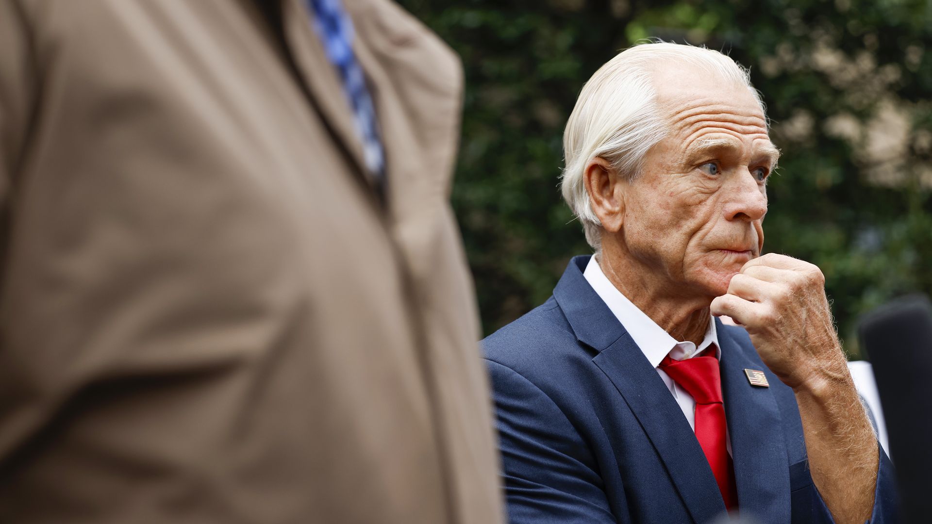 Peter Navarro on the right of the image looking off camera with his hand to his chin. He is wearing a blue suit, white shirt, red tie with an American flag pin. In the foreground, another person's chest is seen in a jacket. 