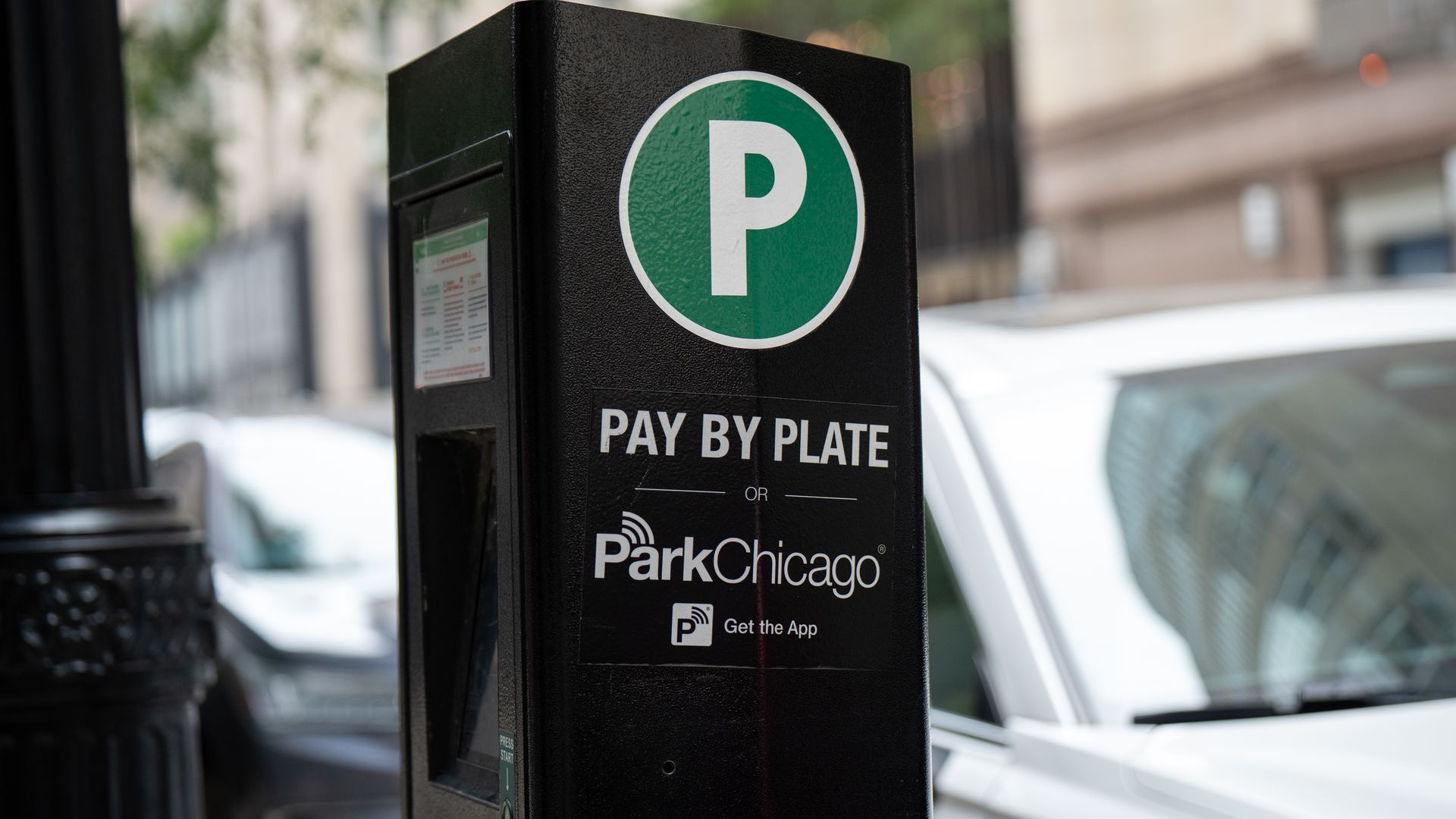 Close-up of a black parking meter with a green circular 'P' logo. The sign shows "PAY BY PLATE" and "ParkChicago"; a blurred city street and cars in the background.
