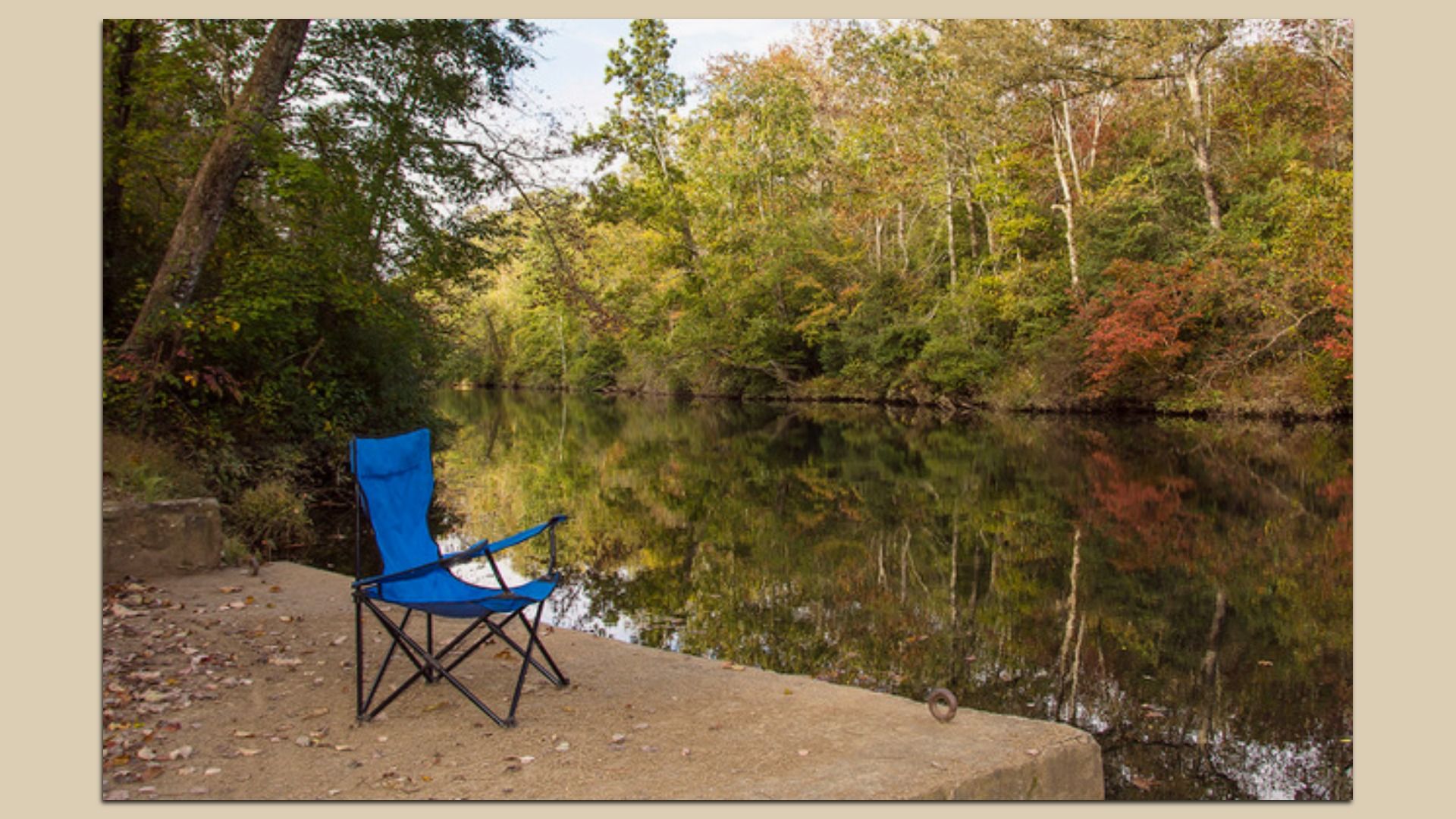 A blue folding camping chair sits on a sandy riverbank by a calm lake, with reflections of green and autumn trees in the water and fallen leaves scattered on the ground.