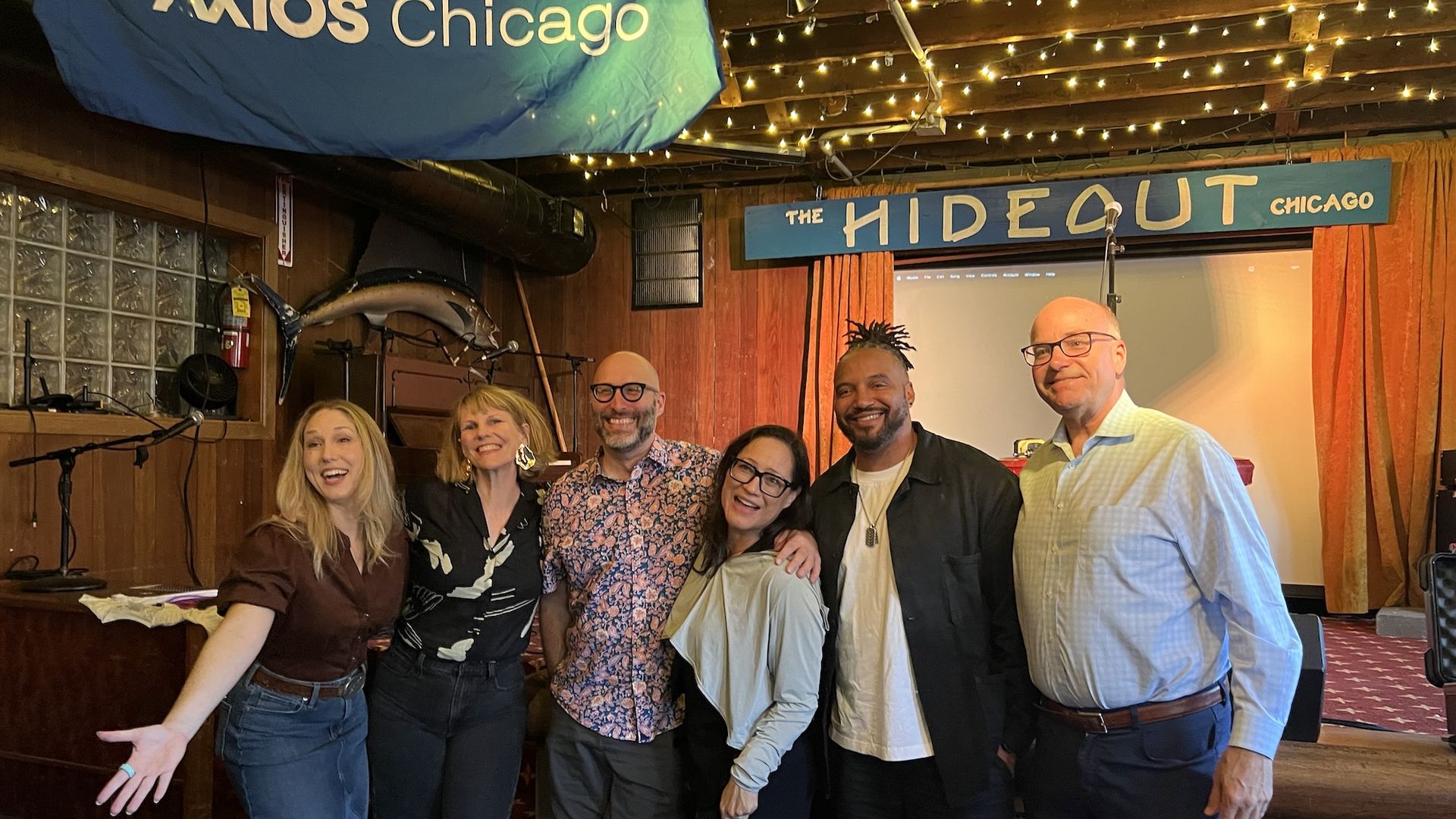 Seven people pose for a group photo in a cozy venue with string lights and wood walls; signs read "THE HIDEOUT CHICAGO" and "AXIOS Chicago" overhead.