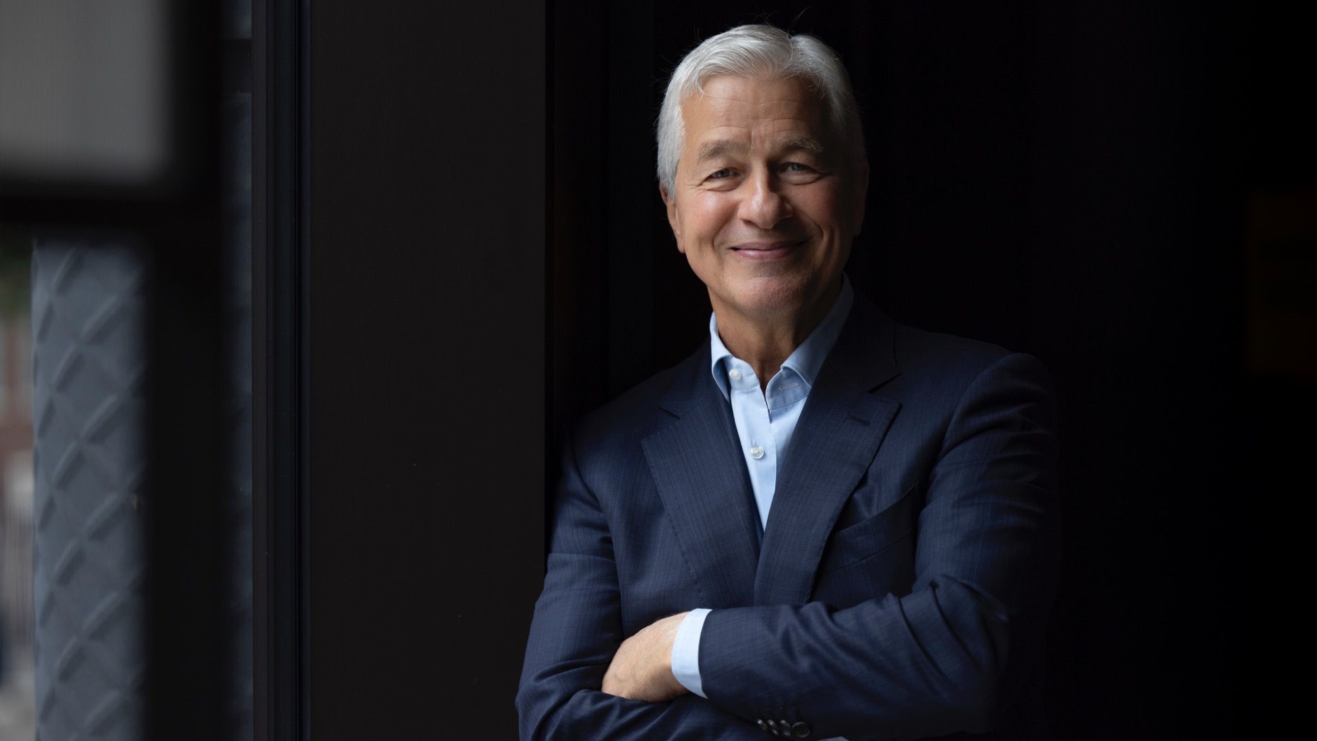 JPMorgan Chase CEO Jamie Dimon with white hair in a dark blue suit and light blue shirt, standing with arms crossed against a dark background near a window.