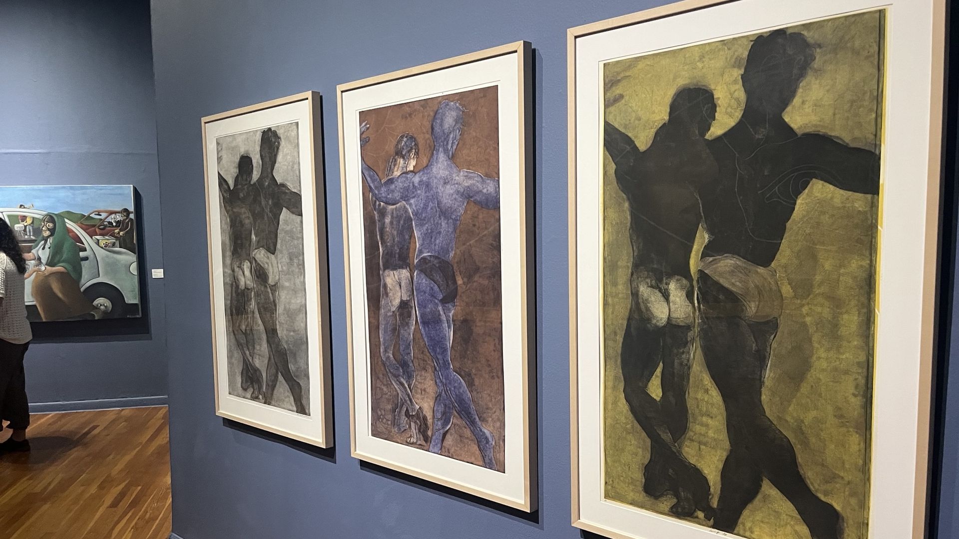 Blue gallery wall with three large framed prints of abstract human silhouettes: left grey, middle blue, right dark on yellow; wooden floor and a partial viewer visible.