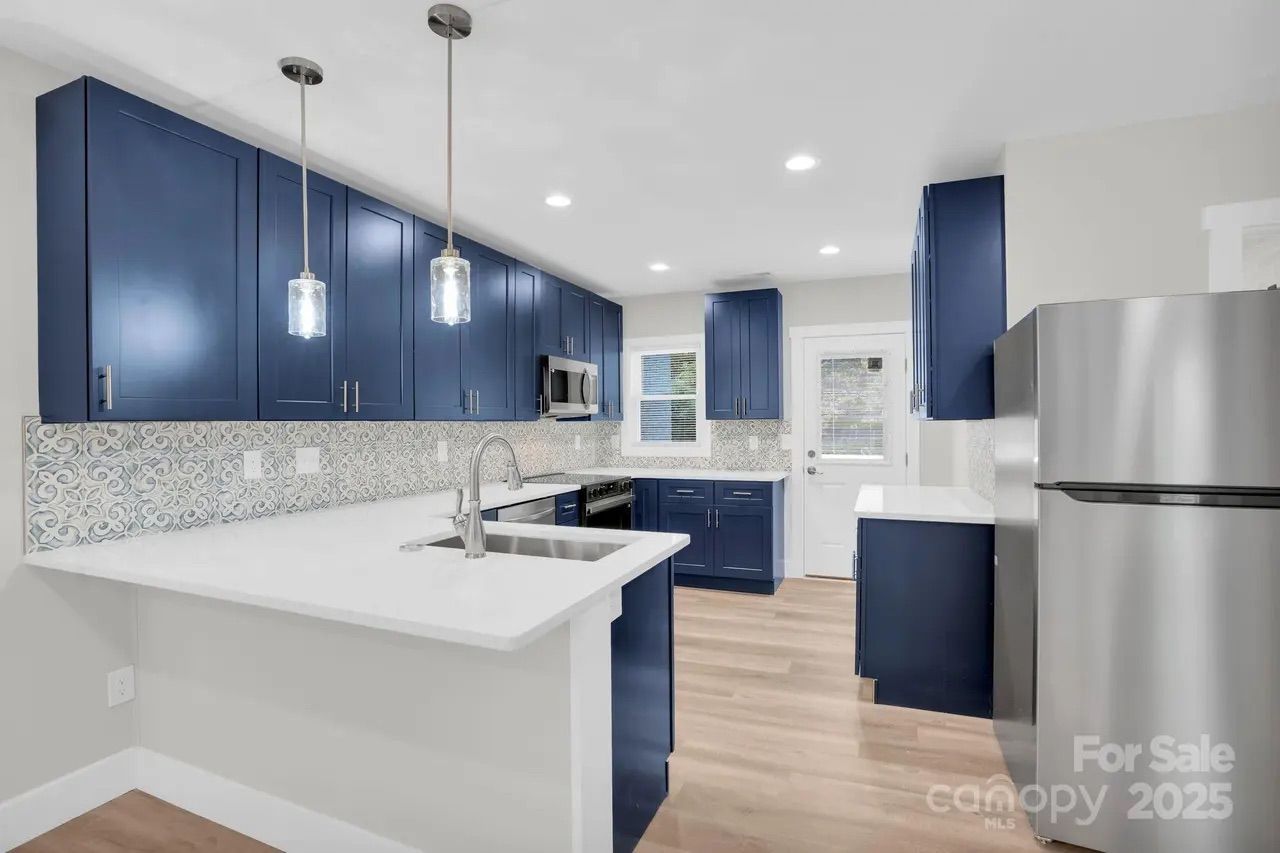 Modern kitchen with navy blue cabinets, white countertops, patterned backsplash, stainless steel appliances, pendant lights, and light wood flooring.