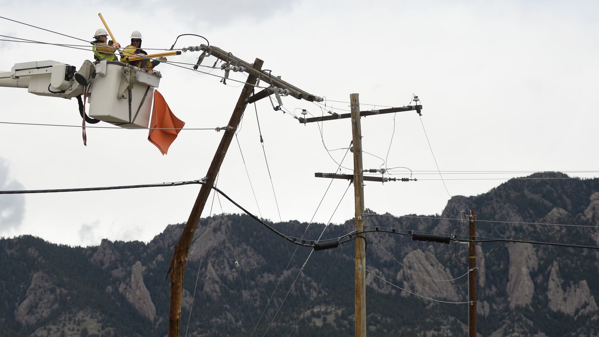 Two utility workers in yellow safety vests and white helmets repairing a leaning power pole from a bucket truck against a mountainous landscape and cloudy sky.
