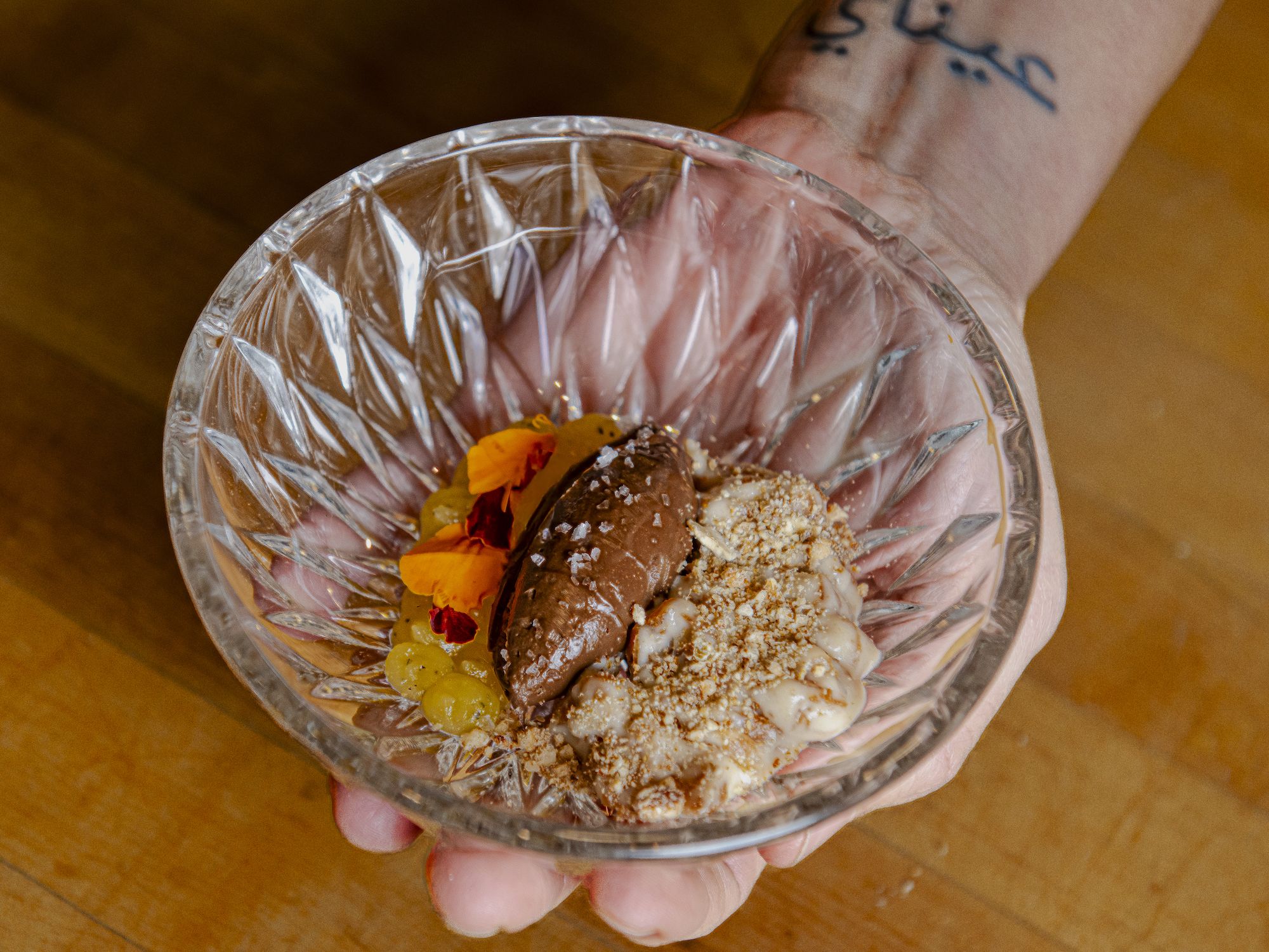 Hand holding a decorative clear glass bowl with a dessert including chocolate mousse, white cream topping, yellow gel dots, orange edible flowers, and brown crumbles on a wooden surface.