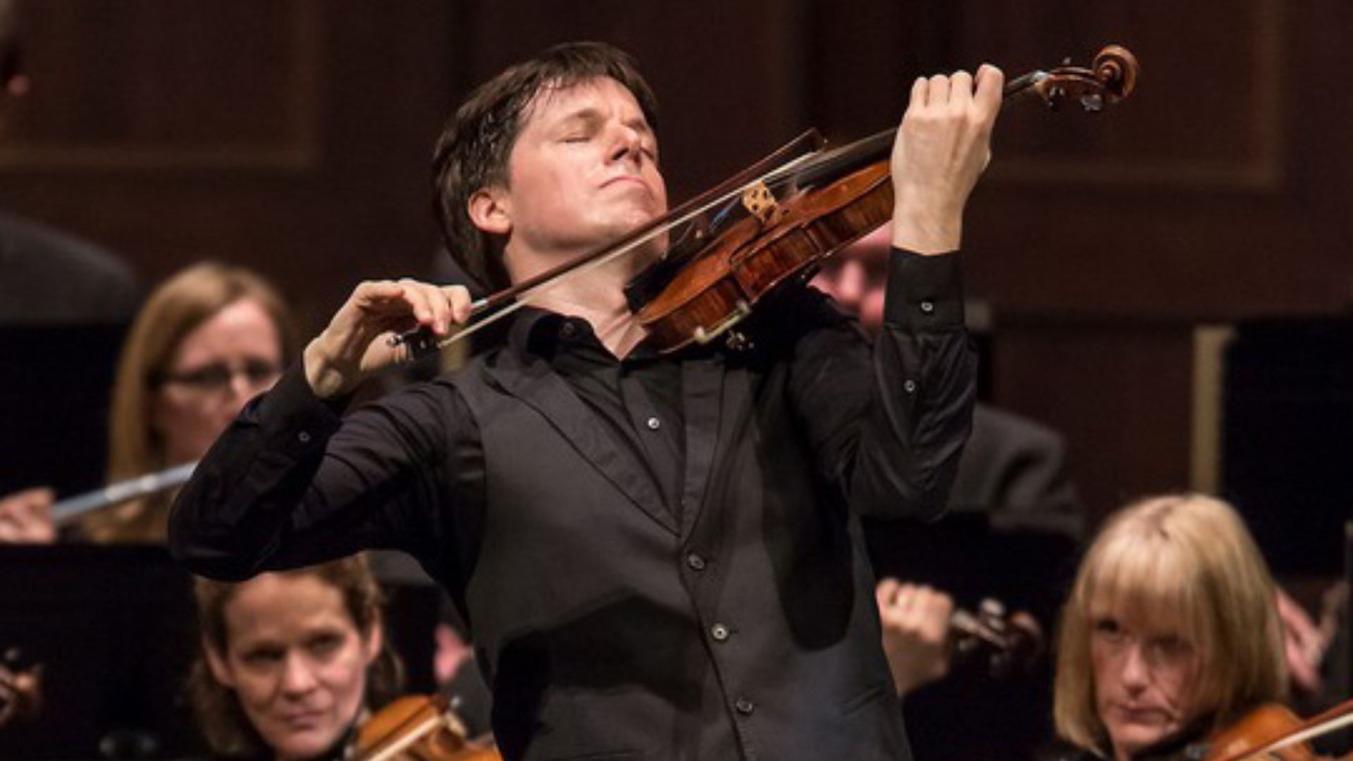 A male violinist in a black shirt and vest plays with eyes closed, bow arcing across a wooden violin, as an orchestra sits in the background on stage.