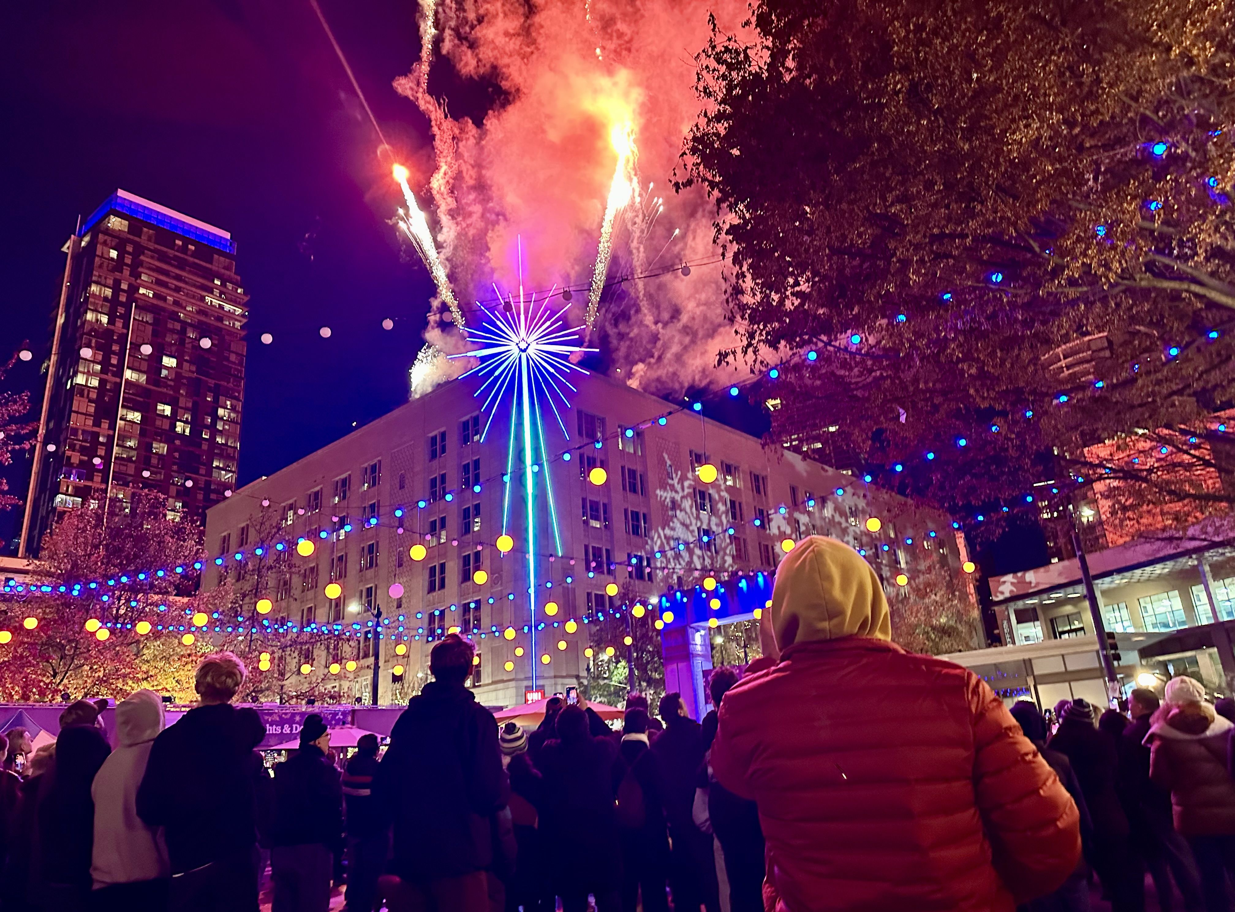 People look at a lit up electric star at the corner of the Macy's building as fireworks shoot overhead. Lights are strung through Westlake Park where people are viewing the spectacle.