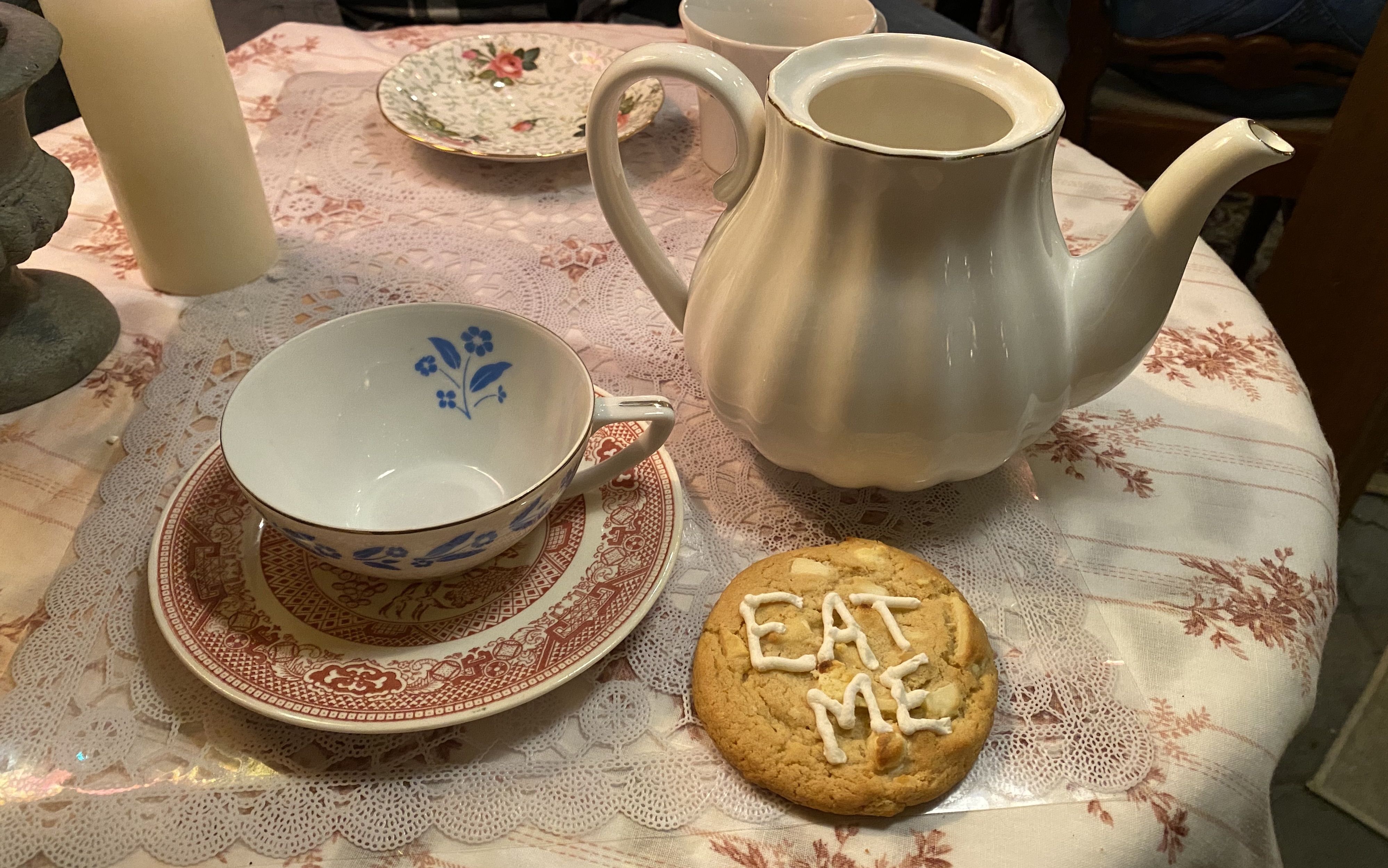A tea set on a table next to a cookie that reads “Eat Me” in frosting