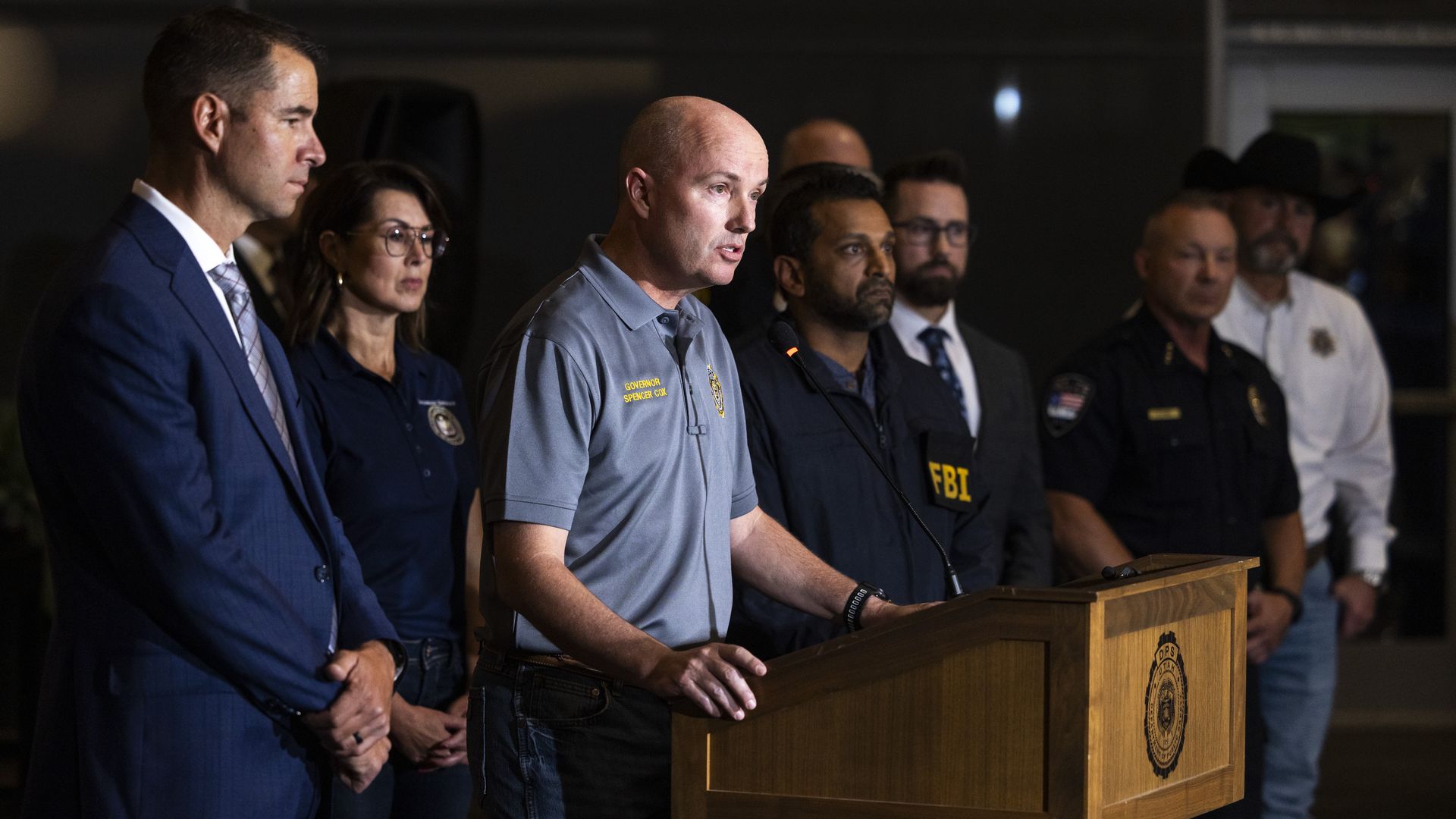 Officials at a press conference: a bald man in a gray polo speaks at a wooden podium, flanked by suited and uniformed colleagues in a dim room.