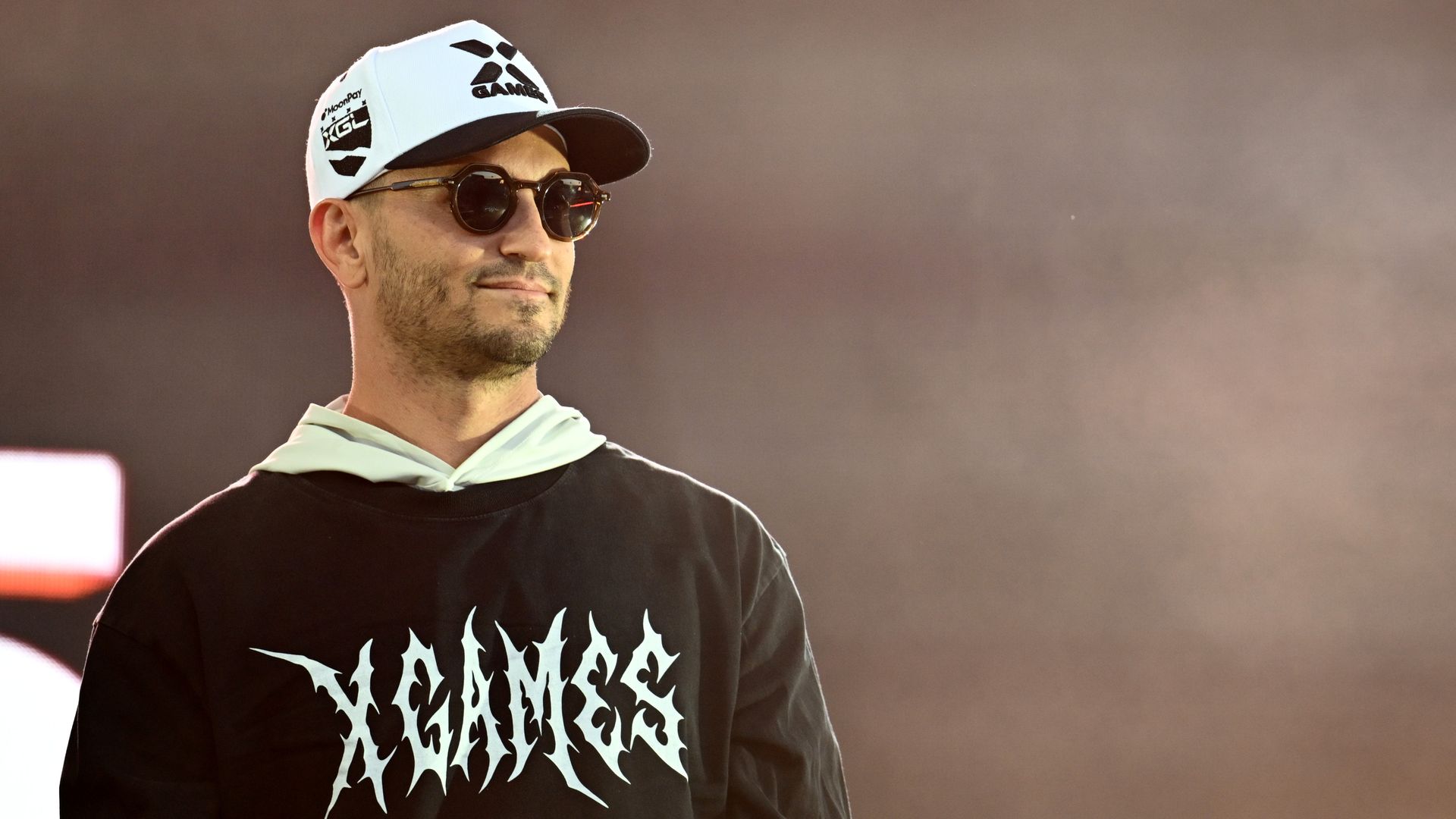 Jeremy Bloom wearing a white cap with black logos and dark sunglasses stands against a hazy brown background; he has a trimmed beard, and wears a black shirt with a white jagged graphic.