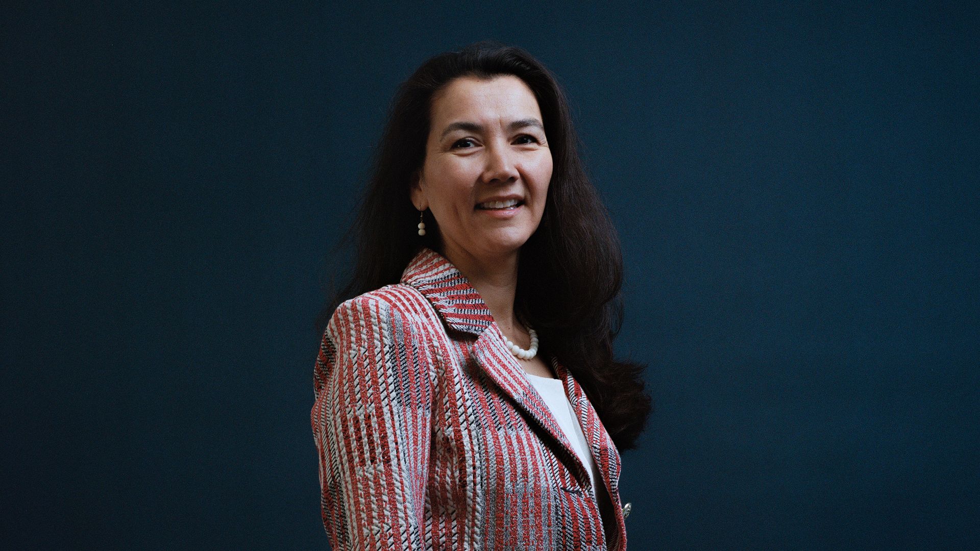 Democrat Mary Peltola, wearing a red and gray jacket, white shirt, pearl necklace and black pants, poses for a photo in front of a dark blue backdrop.