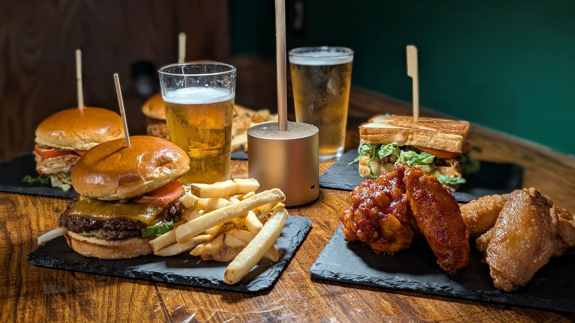 Table with two cheeseburgers with fries, chicken wings, a sandwich with lettuce and tomato, and two glasses of beer under a small lamp on a wooden surface.