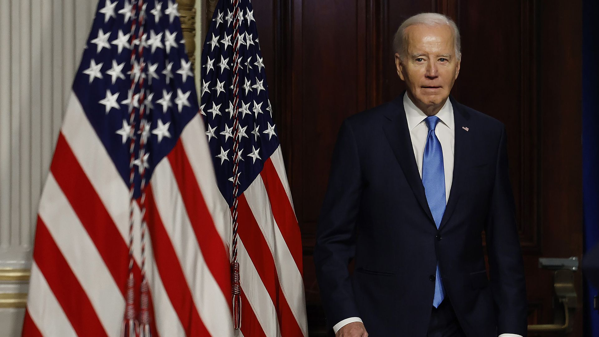 President Biden, in a dark suit, white shirt and blue tie, stands next to several American flags.