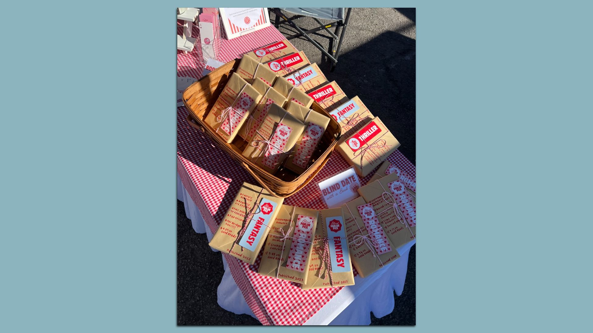 Outdoor market table with a wicker basket of brown gift boxes labeled FANTASY and THRILLER on a red-checkered tablecloth; a sign reads "BLIND DATE with a Book".