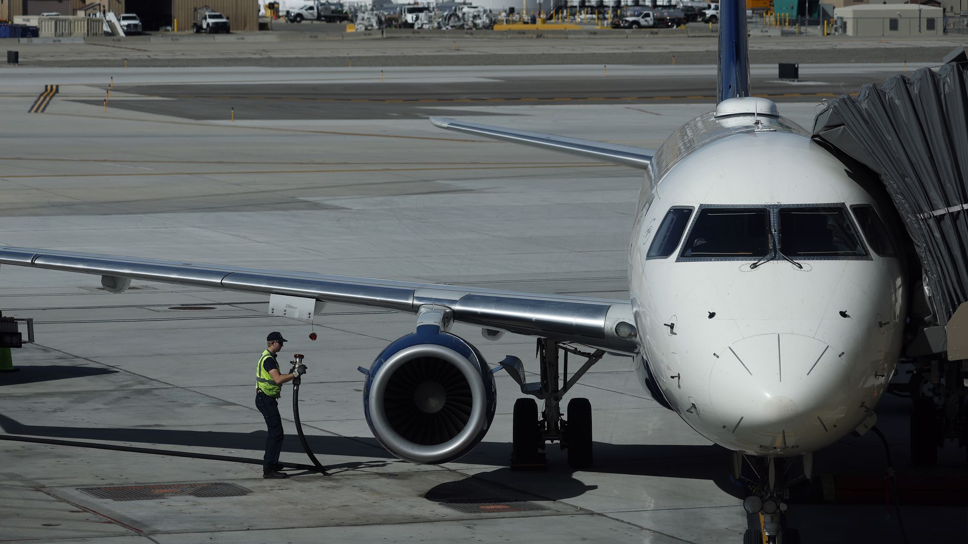 Front view of a passenger jet at a gate with a jet bridge; ground crew in a high-visibility vest handles a hose by the engine, with hangars and distant mountains in the background.