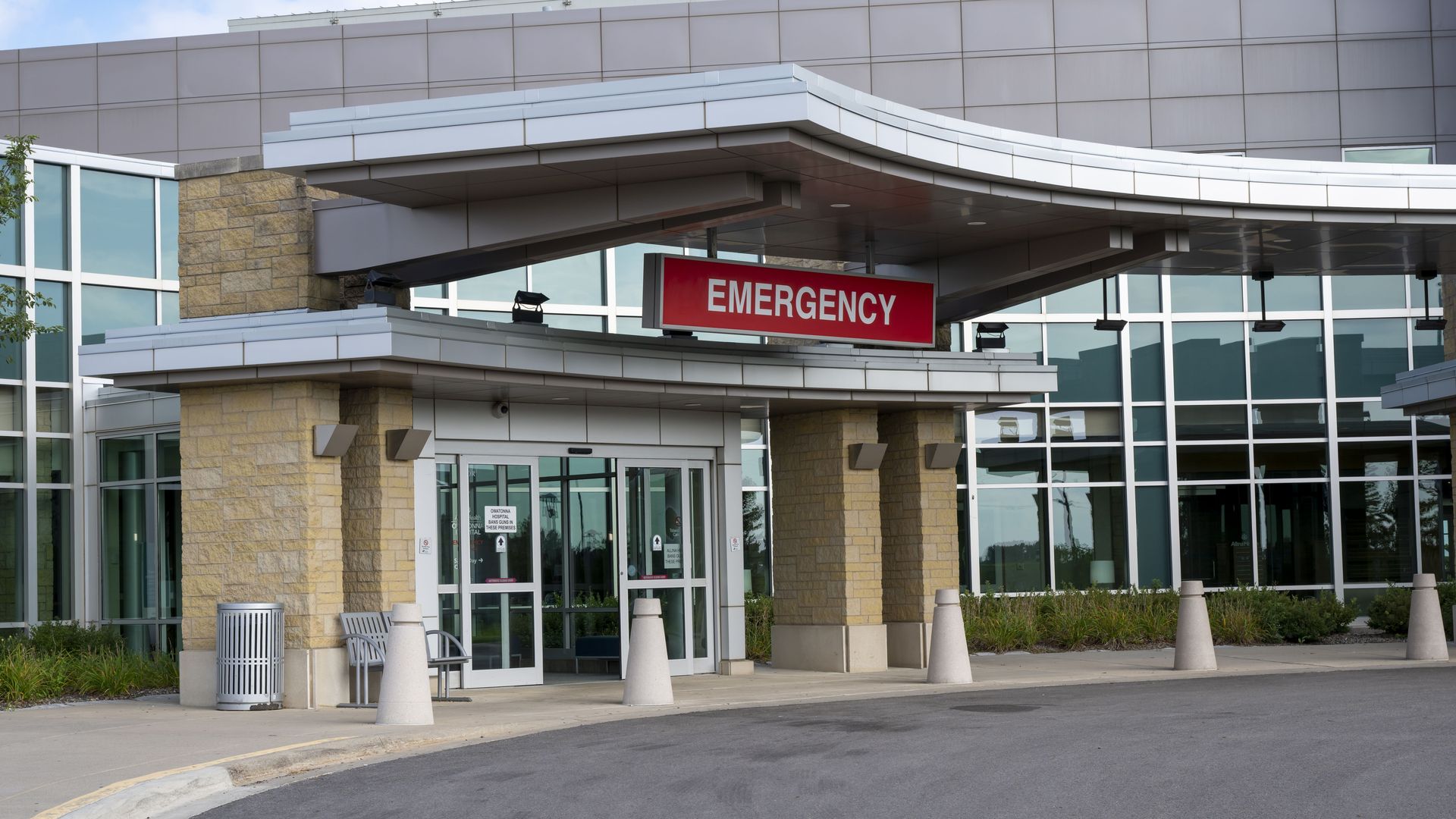 Hospital emergency entrance with a red "EMERGENCY" sign above glass doors, beige stone columns, curved canopy, large glass facade, and a paved driveway with bollards.