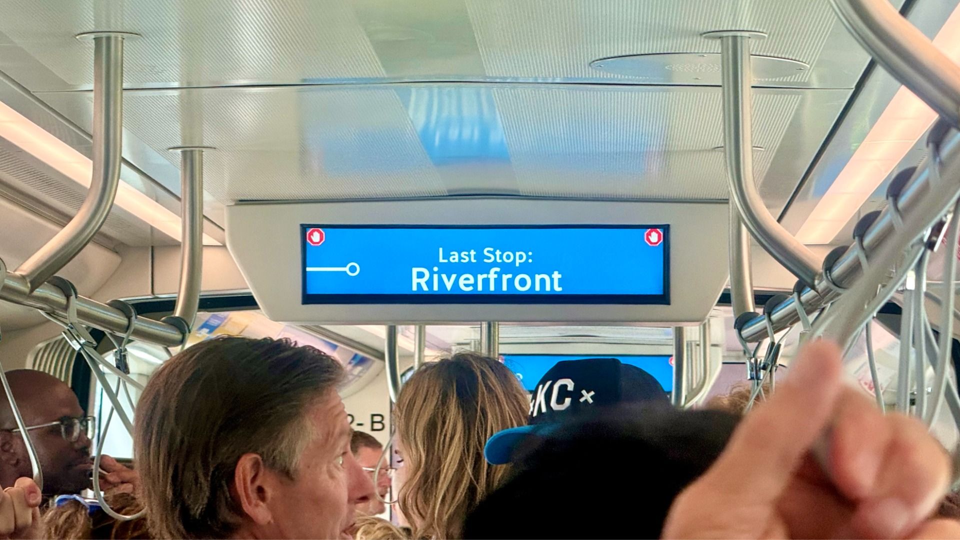 Inside a crowded metro car, a blue digital sign reads "Last Stop: Riverfront" as passengers sit and stand, with curved metal handrails overhead and bright ceiling lights.