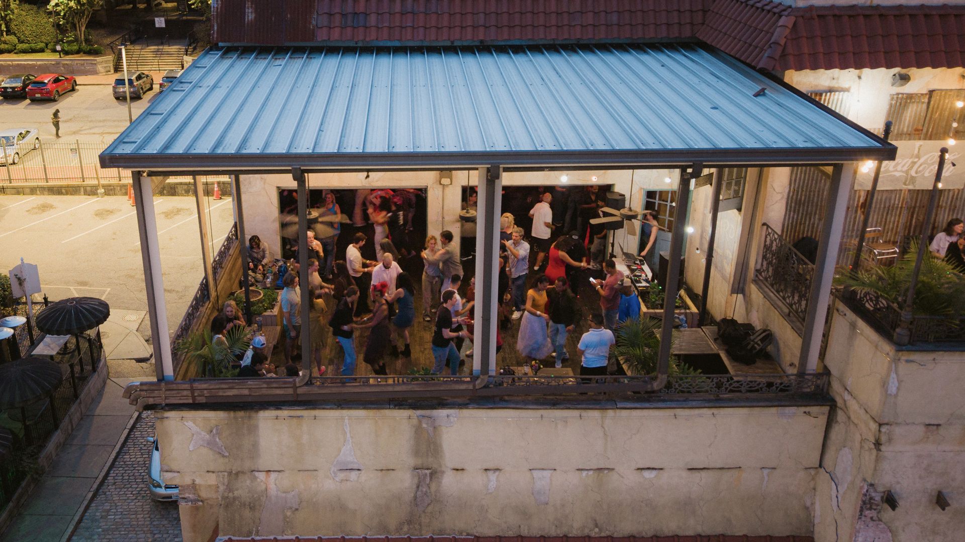 A rooftop party inside a glass-walled venue under a blue-gray metal roof; a crowded dance floor with people mingling under string lights, while cars park along a street below.
