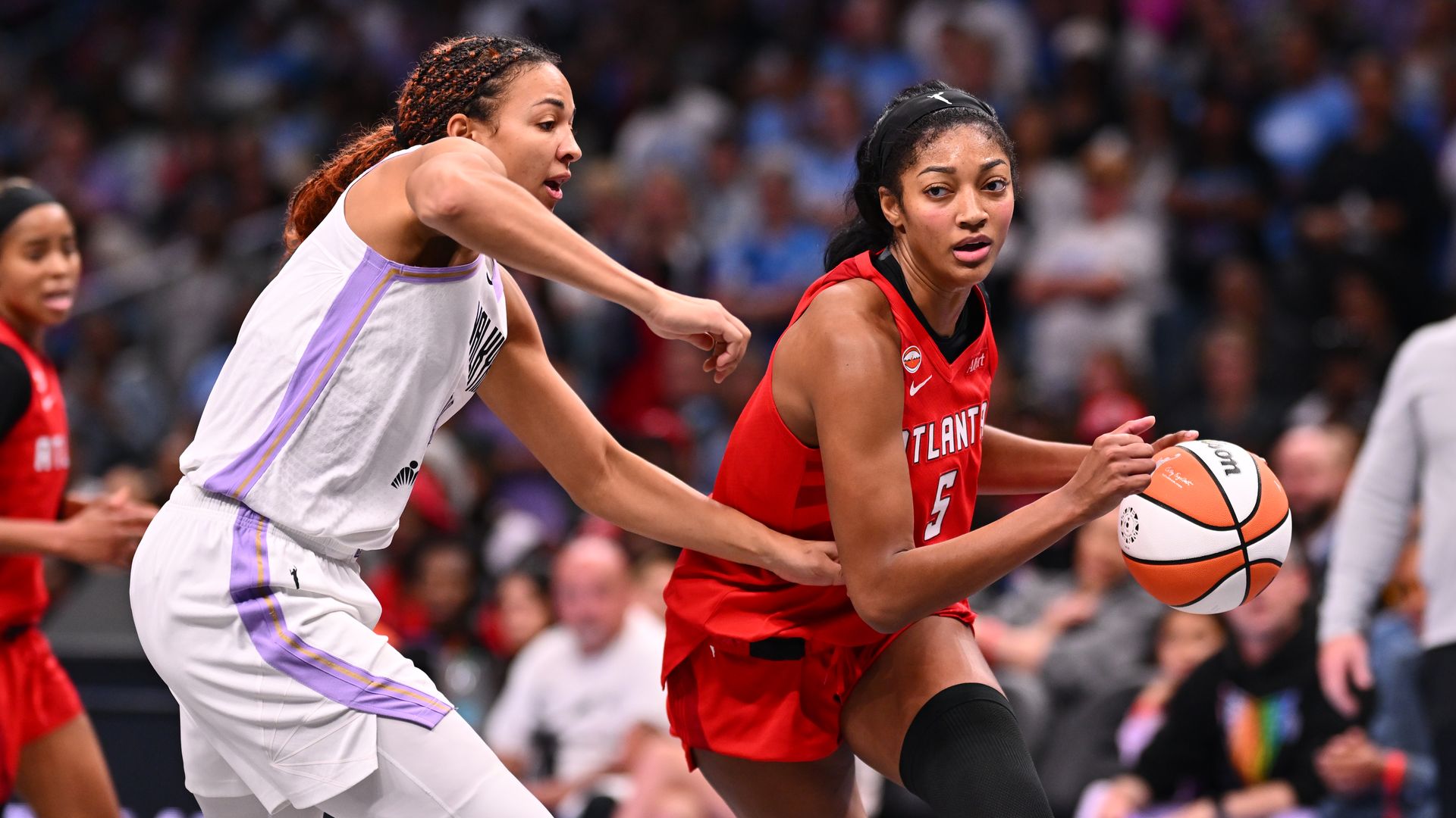 A woman basketball player in a red "ATLANTA" uniform handles the ball while a player in a white uniform attempts to block her.