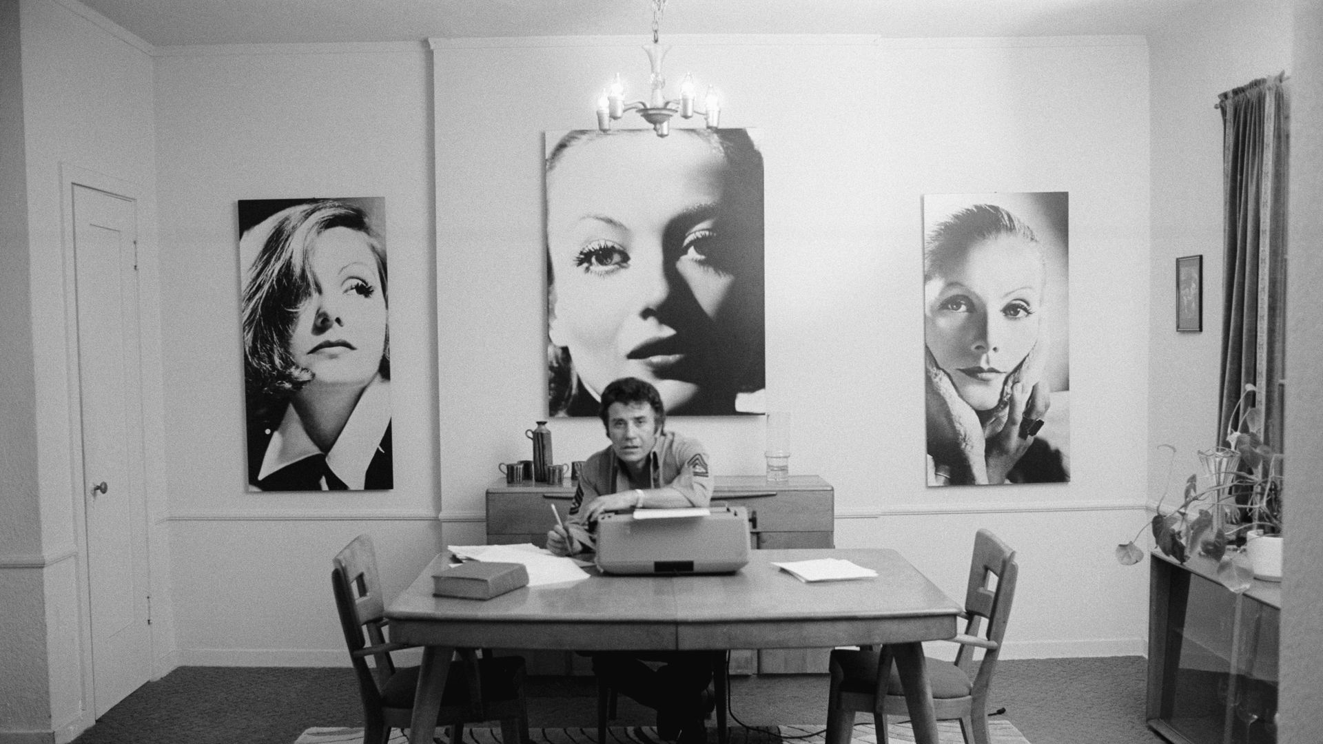 Writer John Rechy at his typewriter in front of large photographs of women.