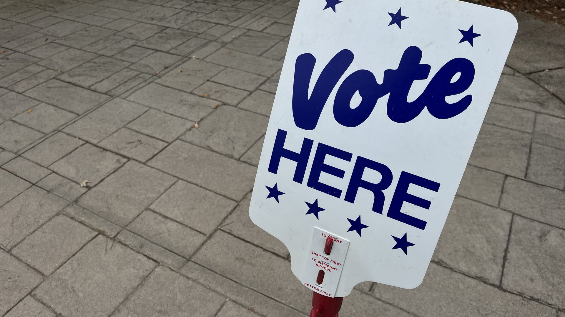 White sign with blue text and stars saying "Vote HERE" on a red stand, placed on a paved sidewalk outdoors with mulch and greenery in the background.