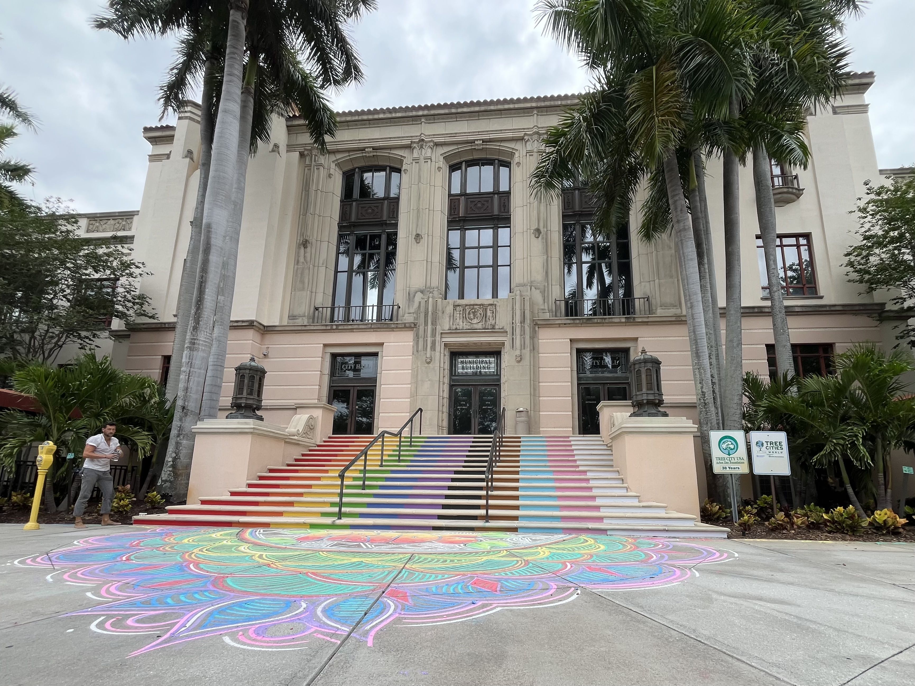Front view of a beige neoclassical building framed by tall palm trees. Rainbow-striped stairs lead to glass doors; a large colorful chalk circle covers the pavement.