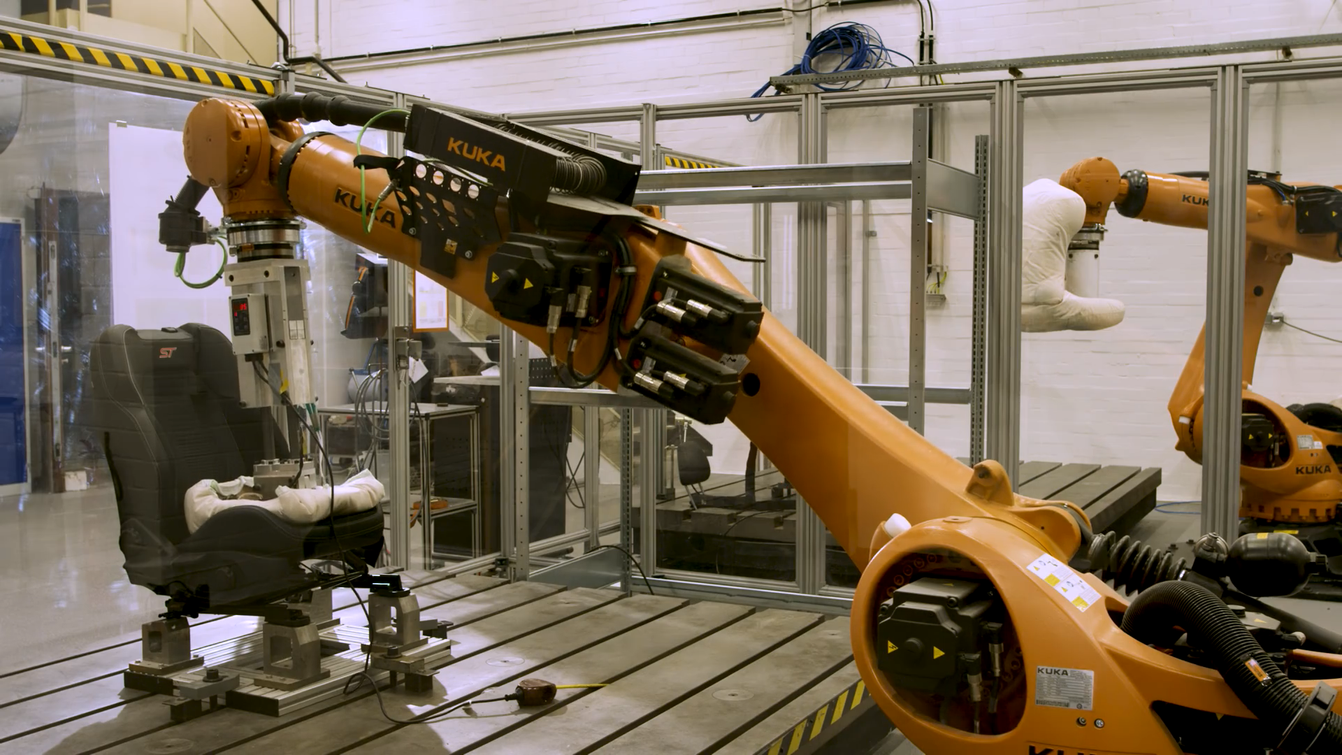 Photo of a robot arm pressing a white pad into the seat of a car in a factory setting.
