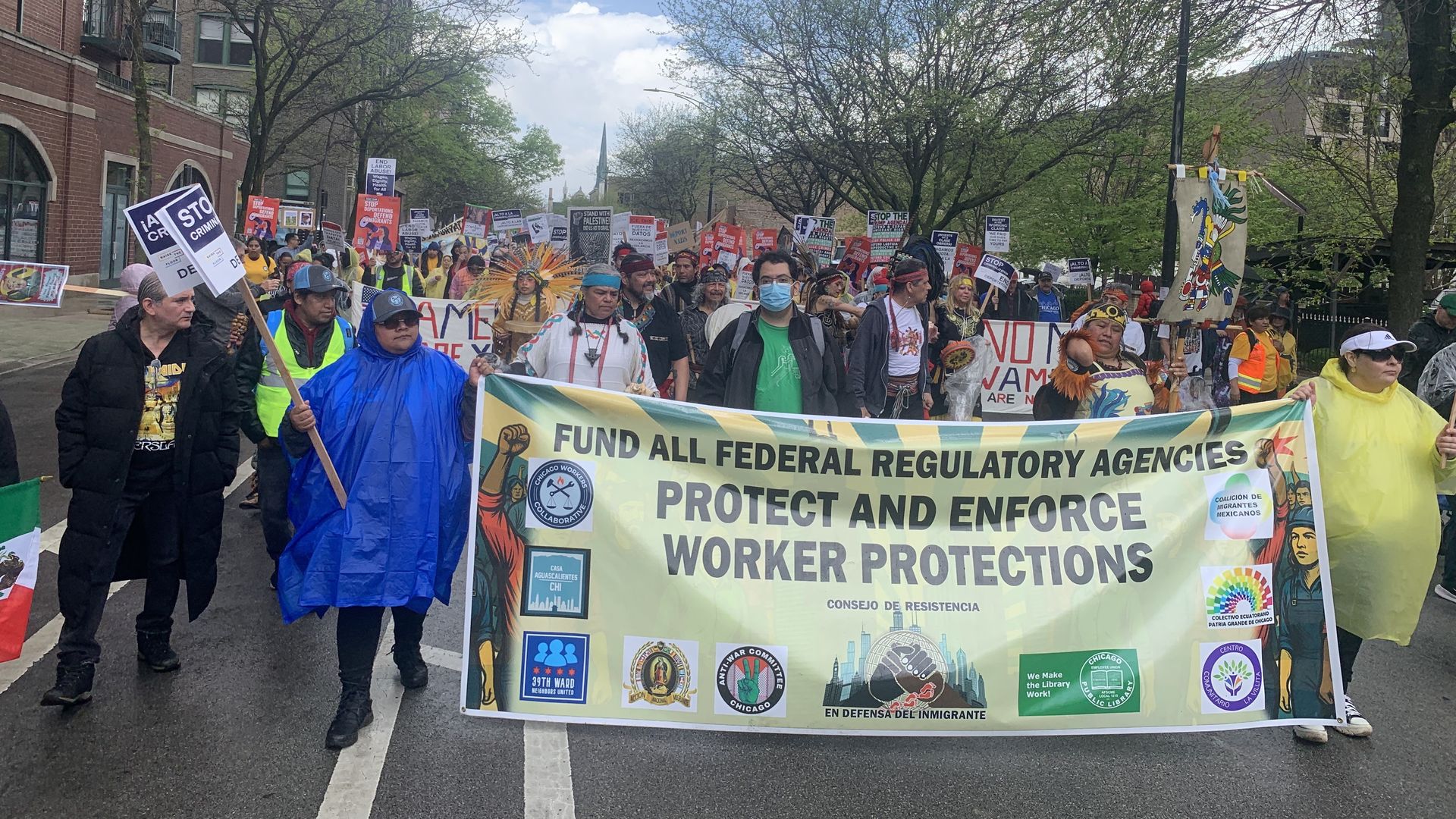Crowd of protesters marches along a city street under a partly cloudy sky. A large banner reads "FUND ALL FEDERAL REGULATORY AGENCIES PROTECT AND ENFORCE WORKER PROTECTIONS" with colorful signs and diverse participants.