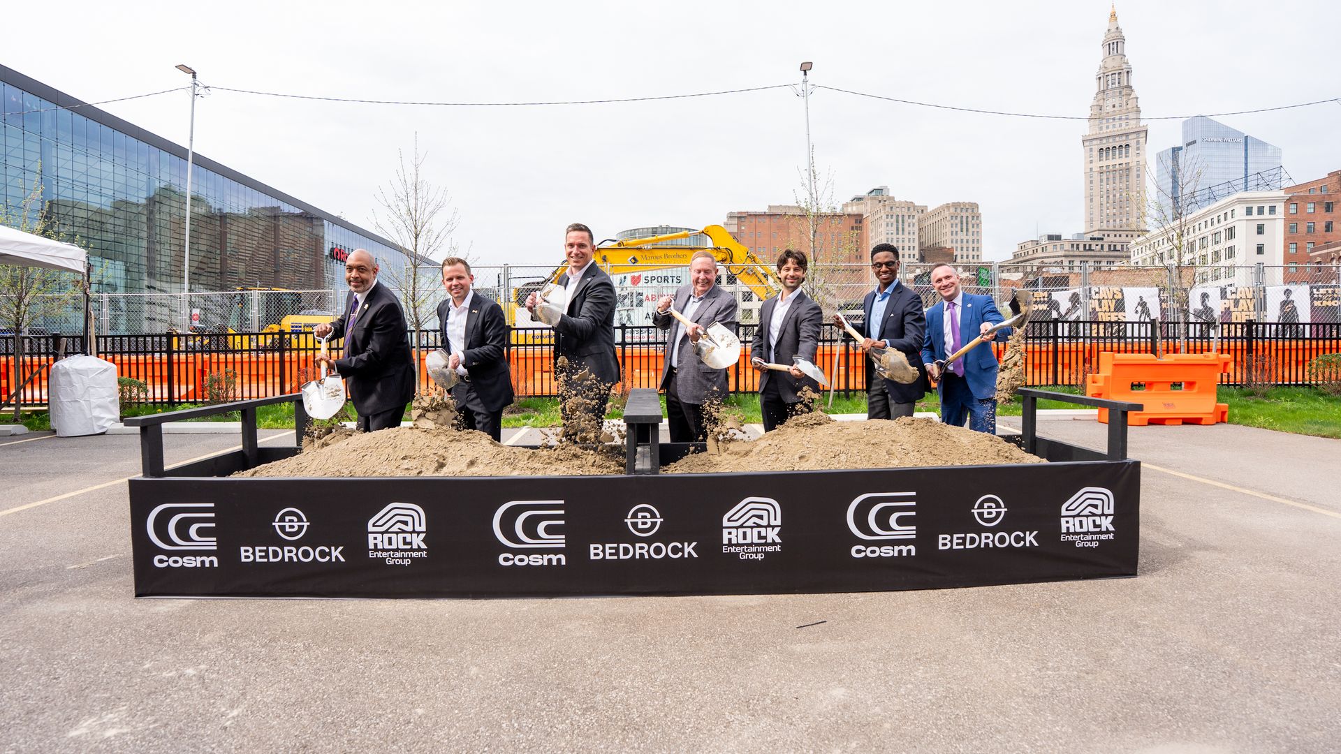 Several men in suits shovel dirt at a groundbreaking ceremony, standing behind a barrier with Cosm, Bedrock, and Rock Entertainment logos. A yellow excavator and city skyline are in the background.