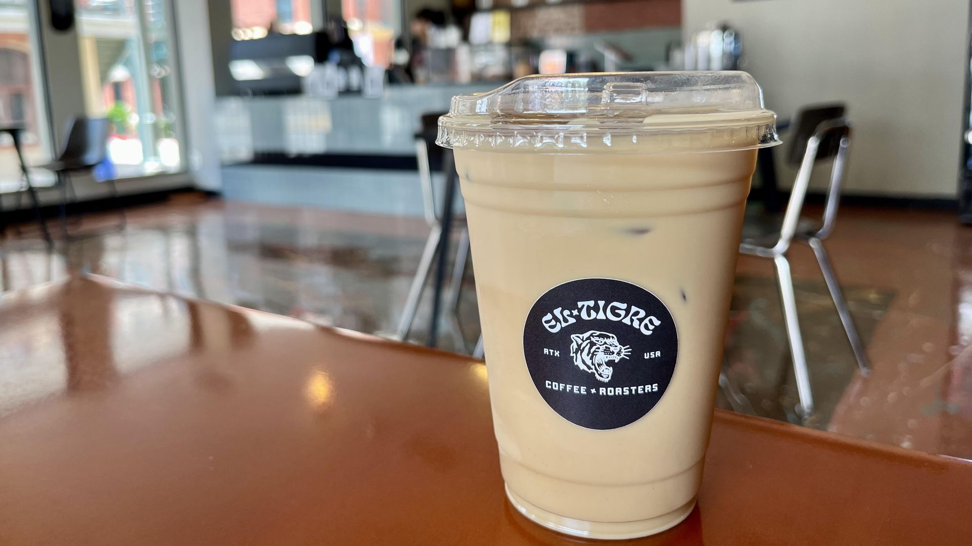 An iced latte with a black "El Tigre" sticker on it sits on a wooden table with a coffee counter blurred in the background.