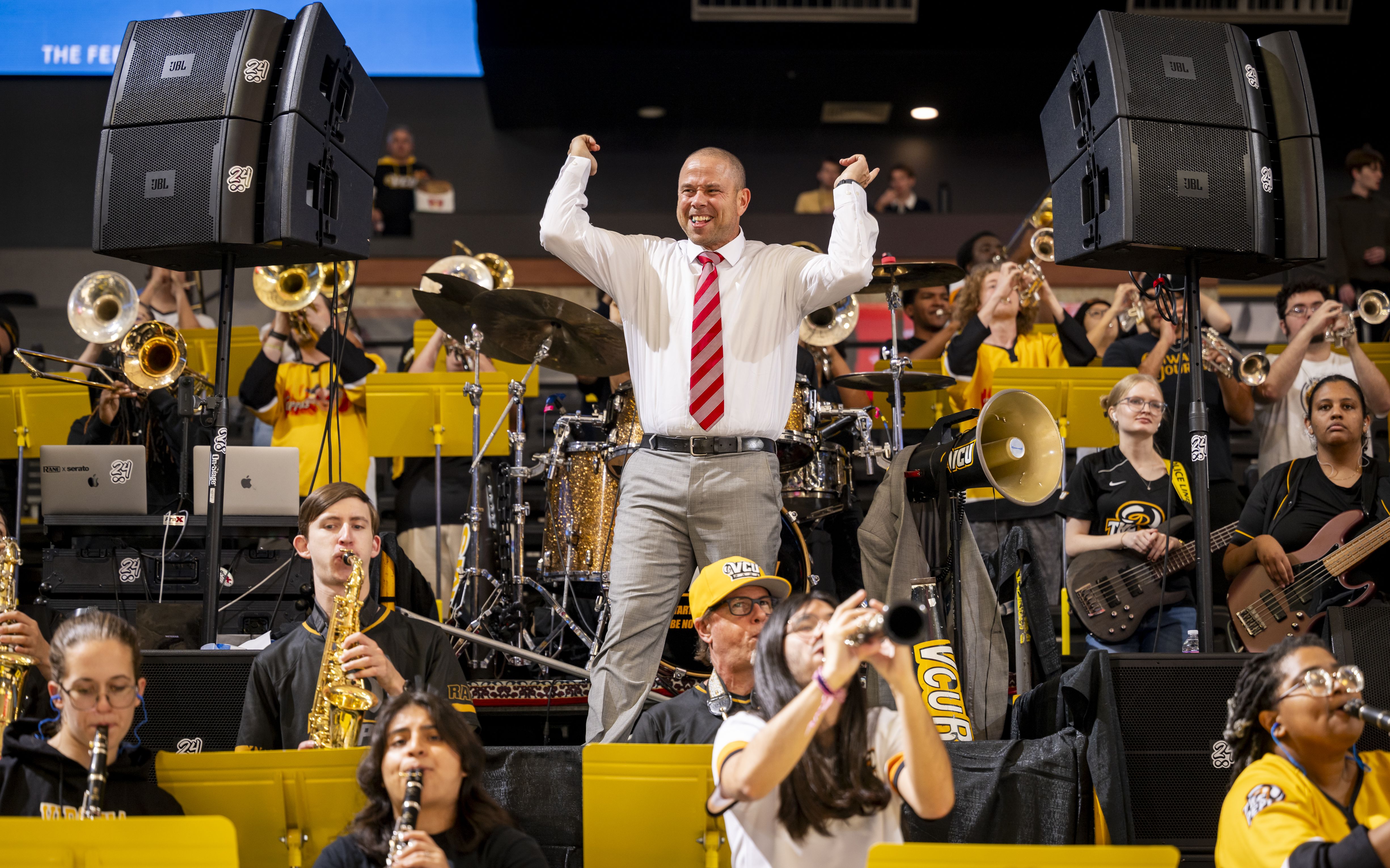 A man in a white shirt and red striped tie conducts a lively band with brass, woodwind, and percussion instruments, all wearing black and yellow clothing, performing indoors with speakers nearby.