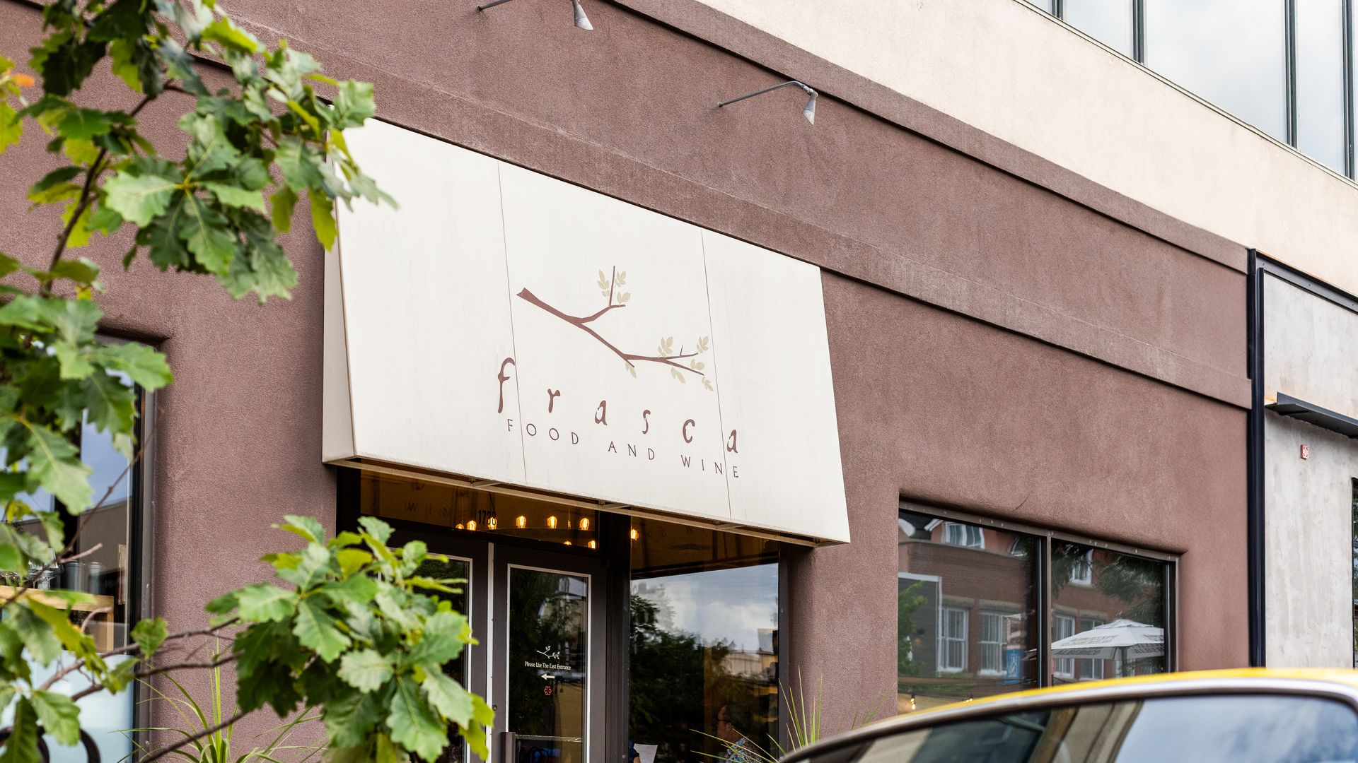 Exterior of Frasca Food and Wine restaurant with a beige awning showing a tree branch logo, brown building facade, colorful flower boxes, and partially visible car in the foreground.