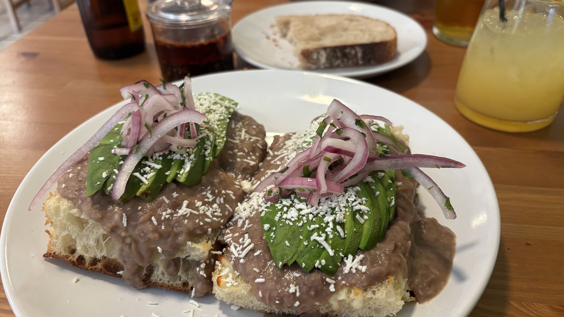 Two slices of toast topped with avocado on a bean spread, shredded cheese, and thin red onion curls, garnished with herbs, on a white plate; background shows a yellow drink and another bread plate.