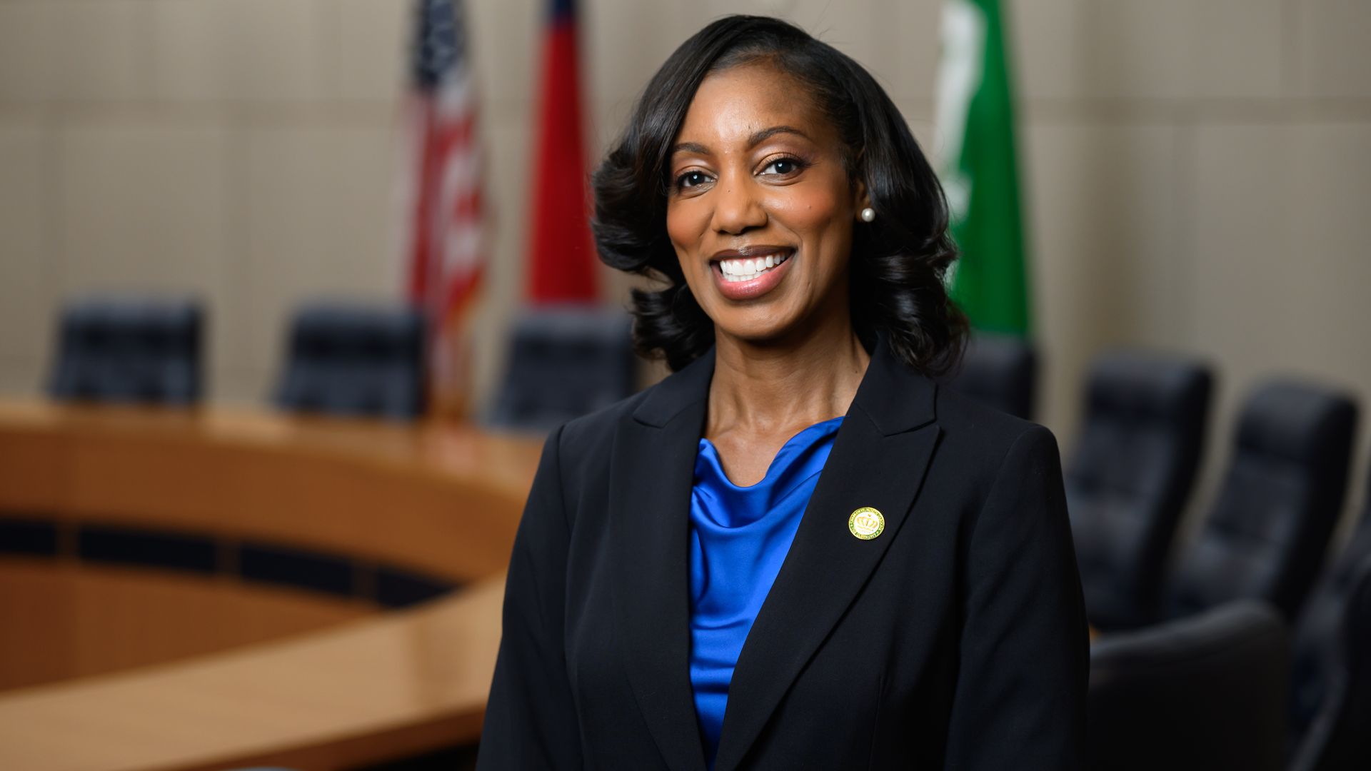 Smiling woman with curled hair wearing a black blazer and blue blouse, standing in a conference room with U.S., red, and green flags in the background.